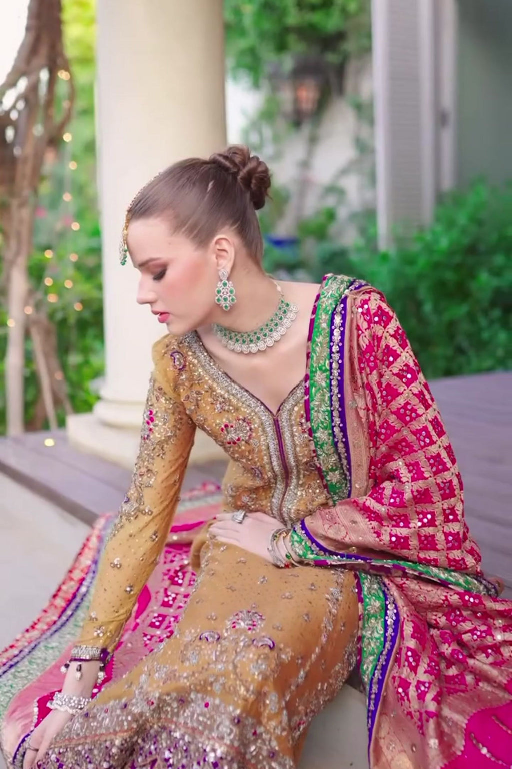 Woman in traditional embroidered outfit sitting outdoors with greenery in the background