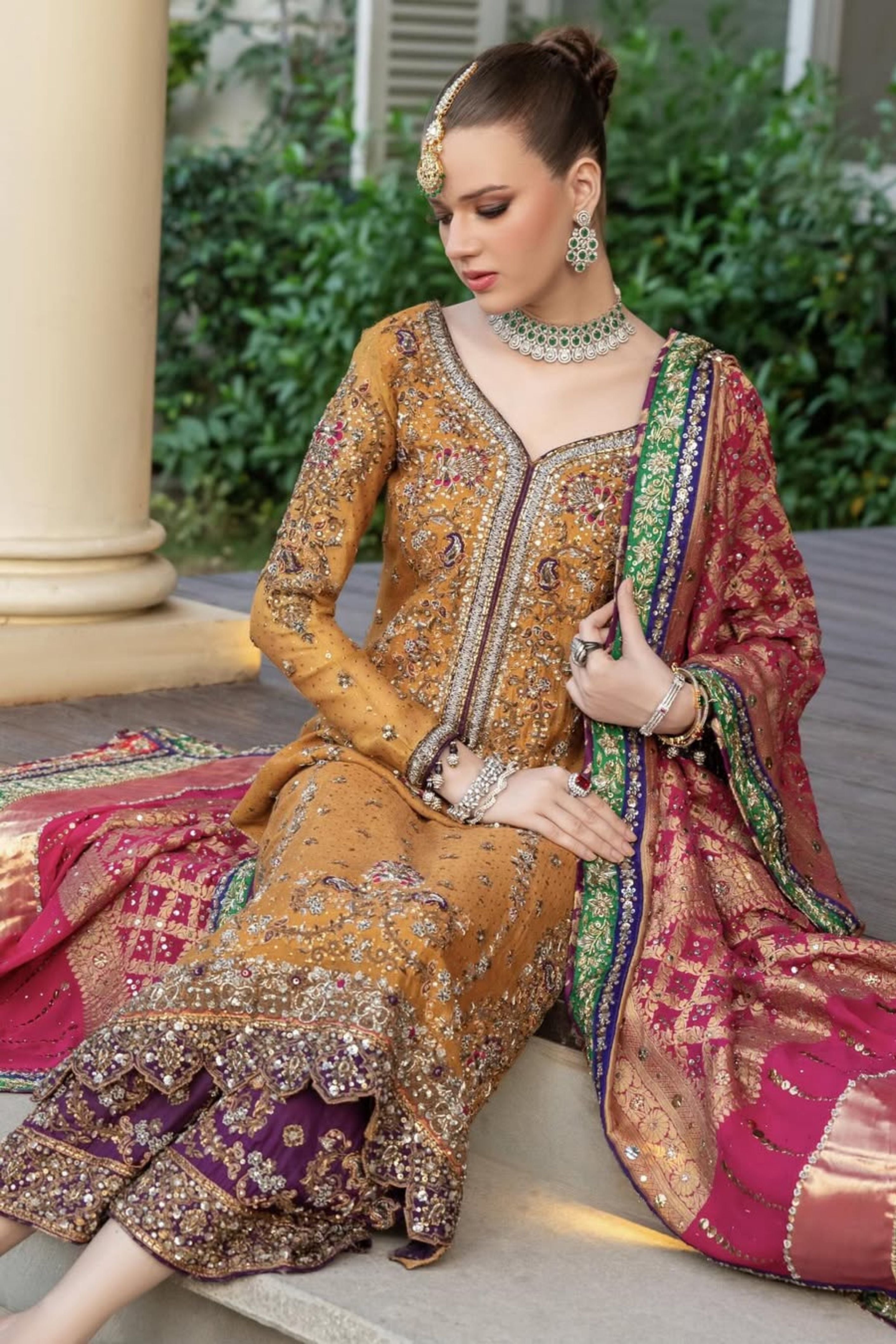 Woman in traditional embroidered gold brown and red color outfit with jewelry, sitting outdoors.