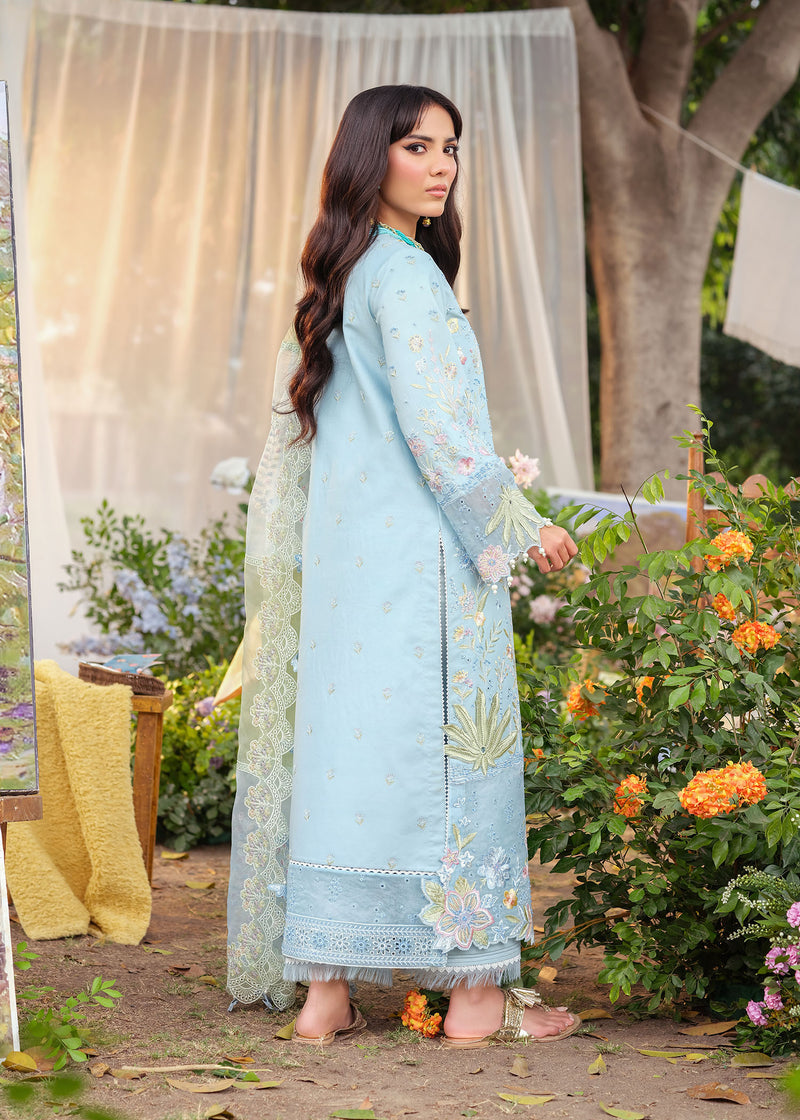 Woman in a light blue embroidered outfit standing outdoors with flowers and a white curtain in the background