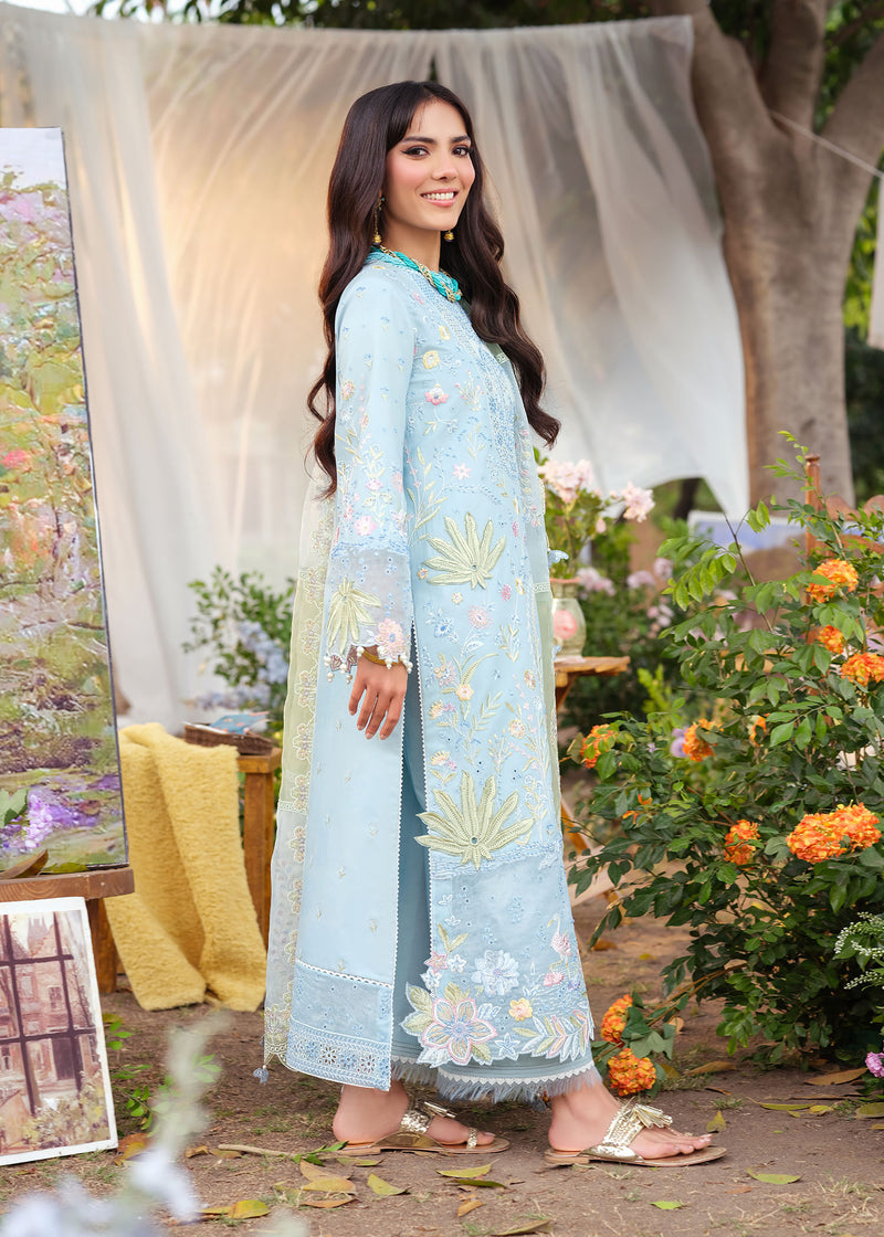 Woman in a light blue embroidered outfit standing outdoors with flowers and a white curtain in the background