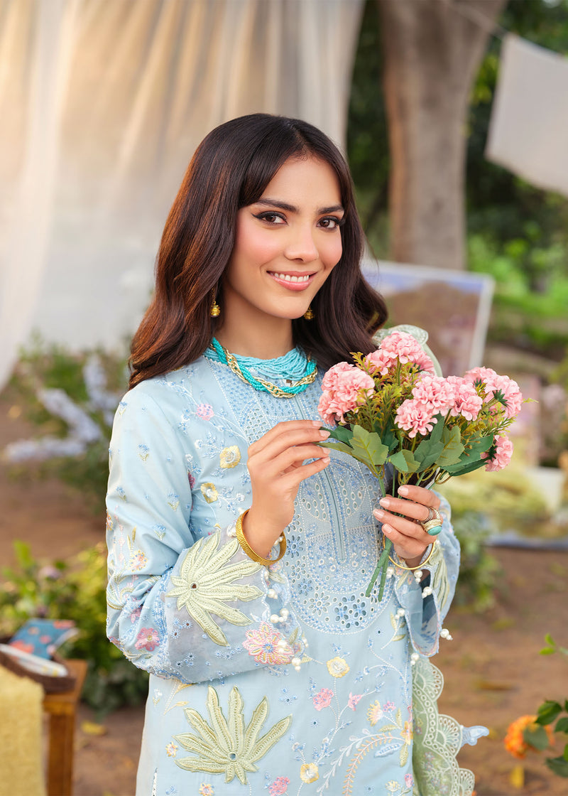 Woman in a light blue embroidered outfit holding pink flowers outdoors.