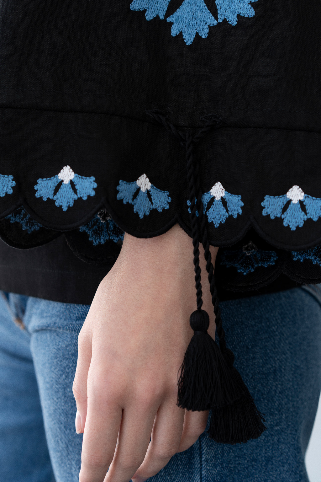 Close-up of a black garment with blue floral patterns and a tassel, held by a person wearing blue jeans.