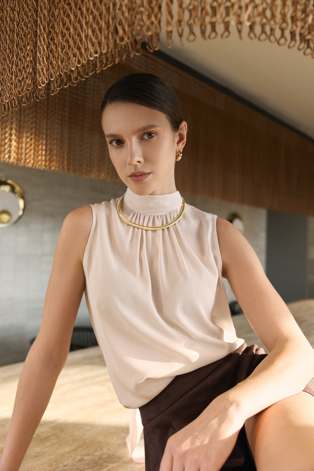 Woman wearing a beige sleeveless top with a gold necklace in an indoor setting