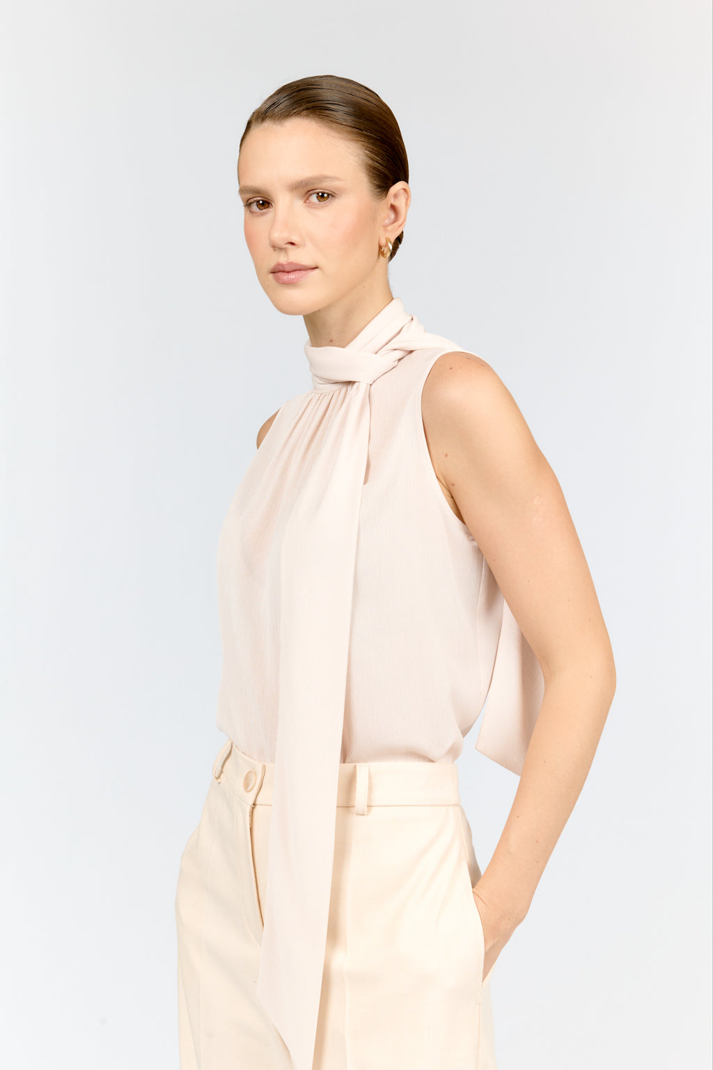 Woman wearing a sleeveless beige blouse with a tie neckline against a light gray background