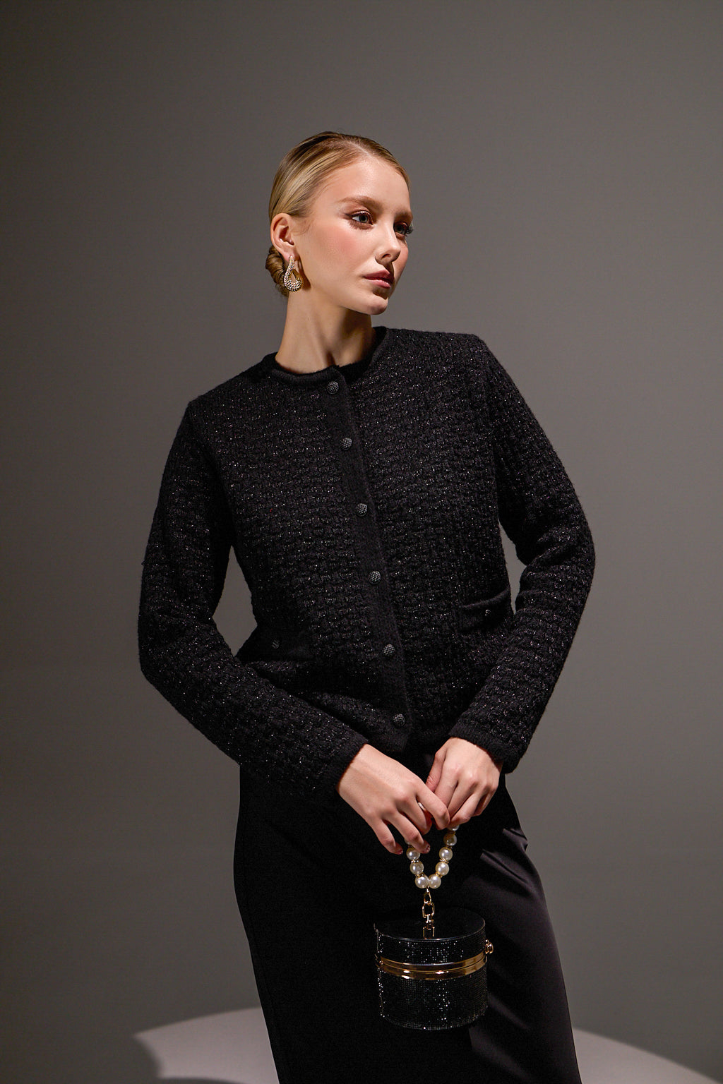 Woman wearing a black textured jacket holding a black handbag against a gray background