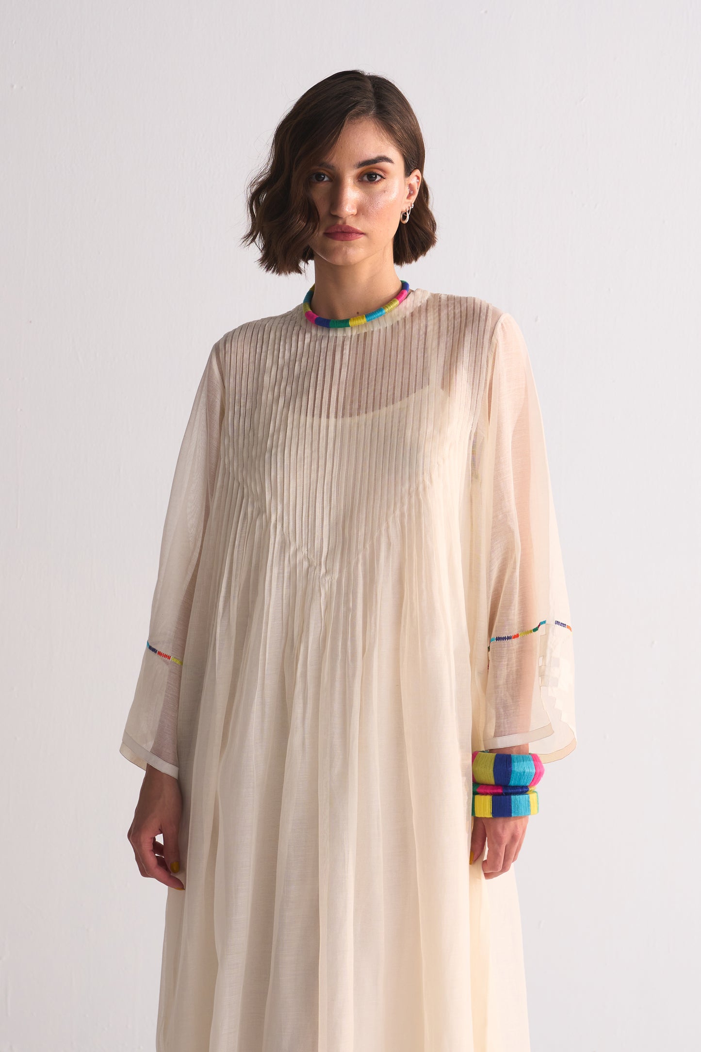 Woman wearing a beige pleated dress with colorful cuffs and neckline against a white background