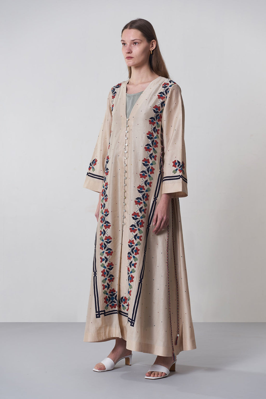 Woman wearing a long, embroidered beige dress with floral patterns on a plain background