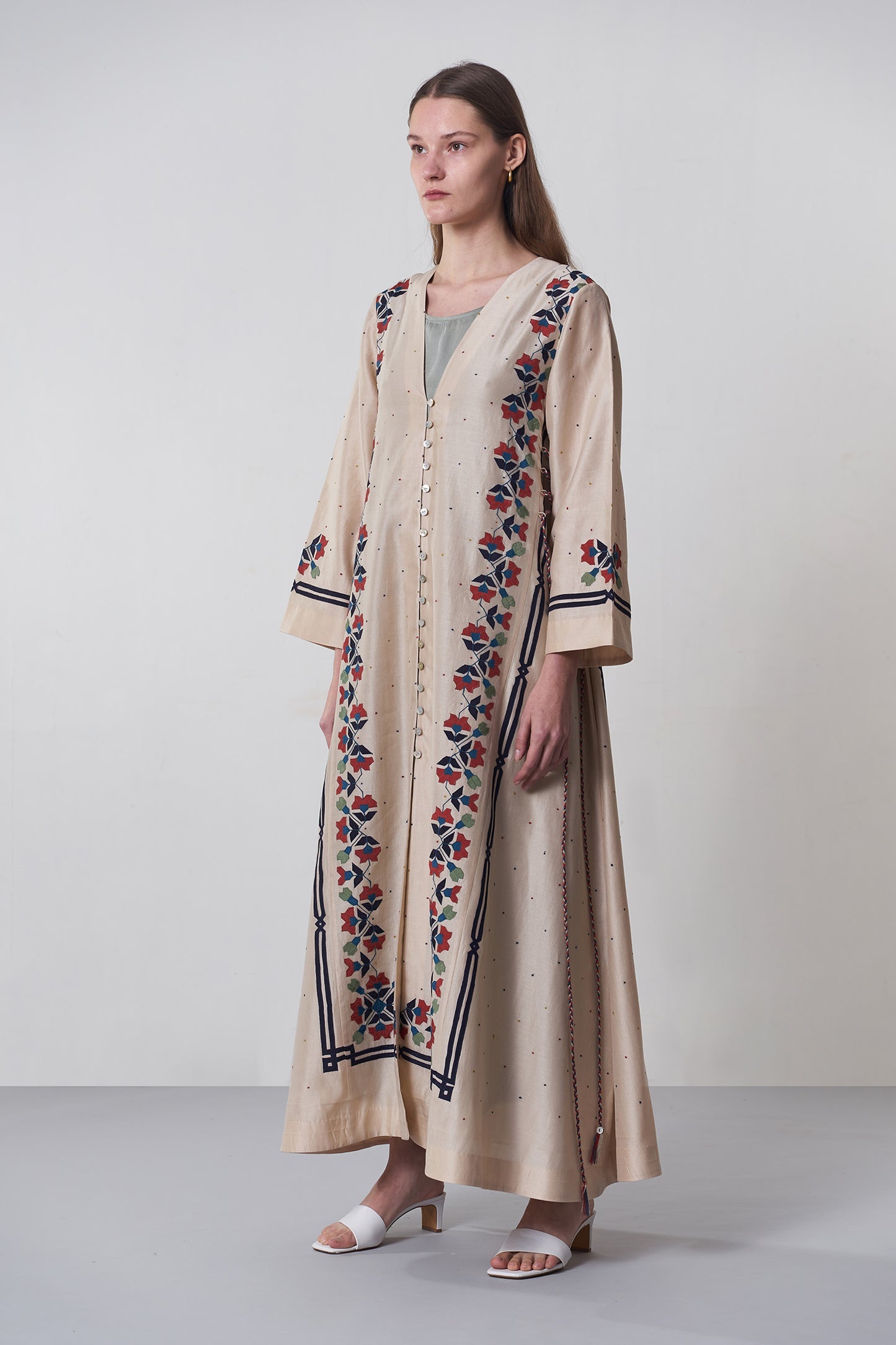 Woman wearing a long, embroidered beige dress with floral patterns on a plain background