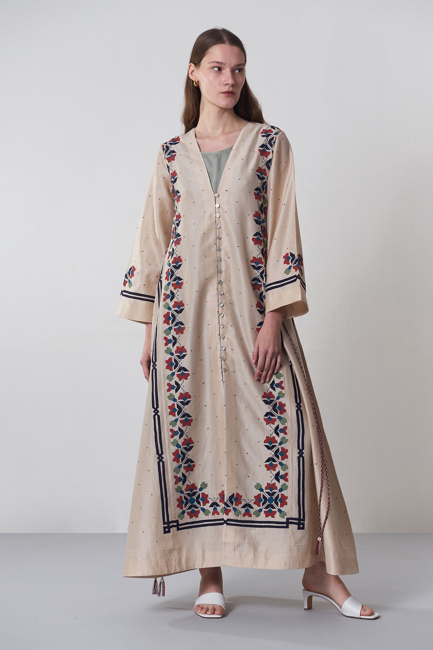 Woman wearing a long beige embroidered dress on a plain background
