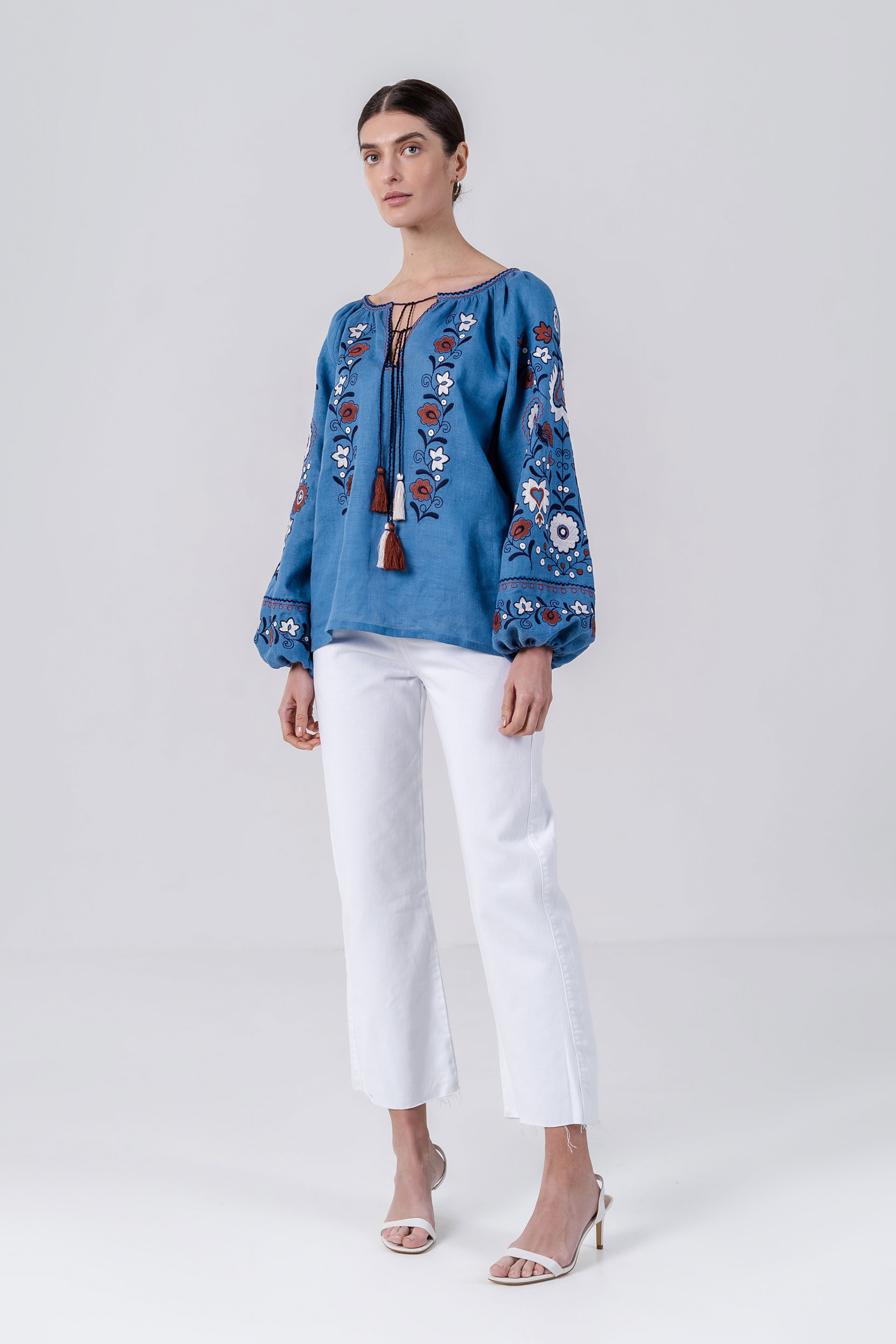 Woman wearing a blue embroidered blouse and white pants on a white background