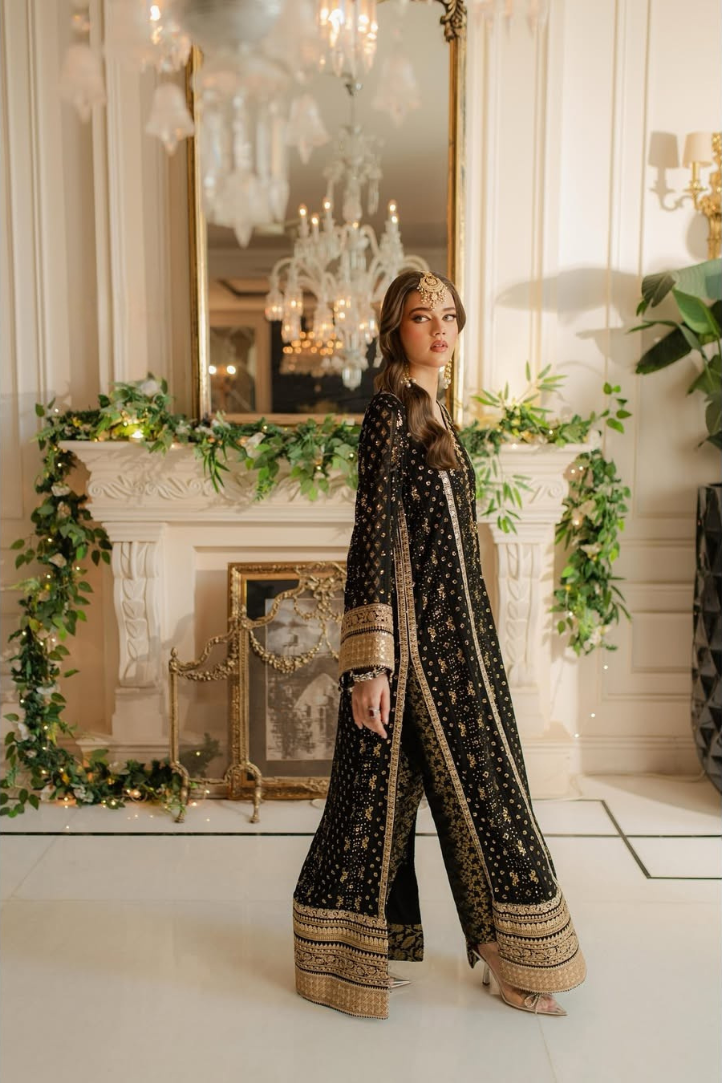 Woman in a black and gold traditional outfit standing in an elegant room with chandeliers and decorative plants.