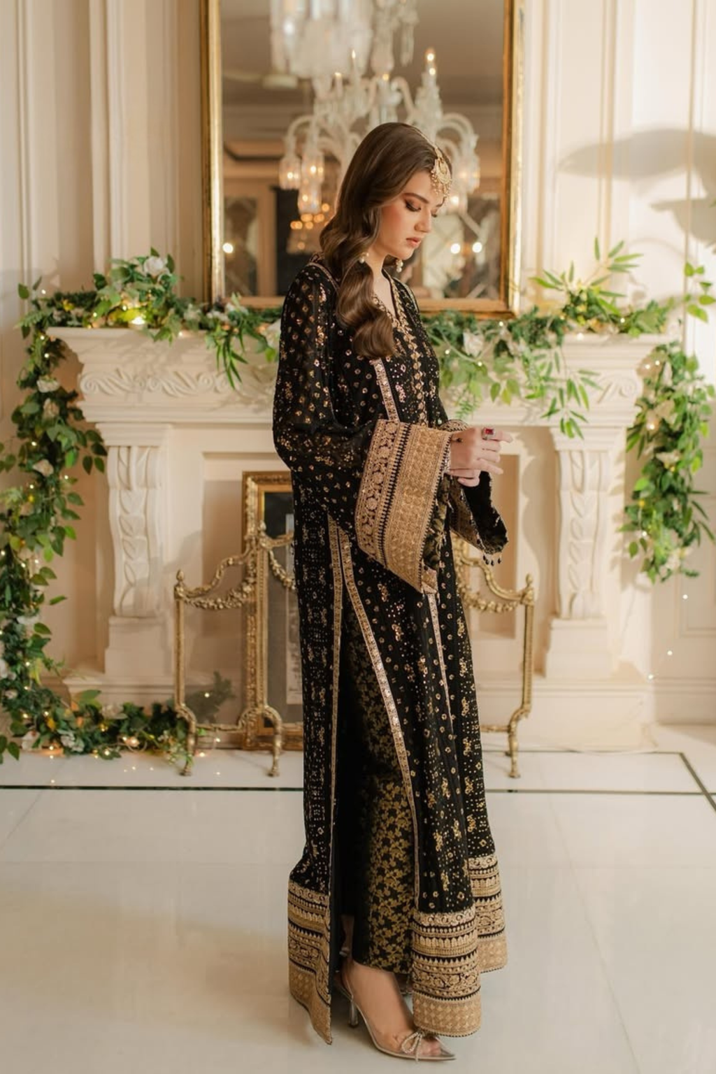 Woman in a black and gold dress standing in a decorated room with greenery and a mirror.