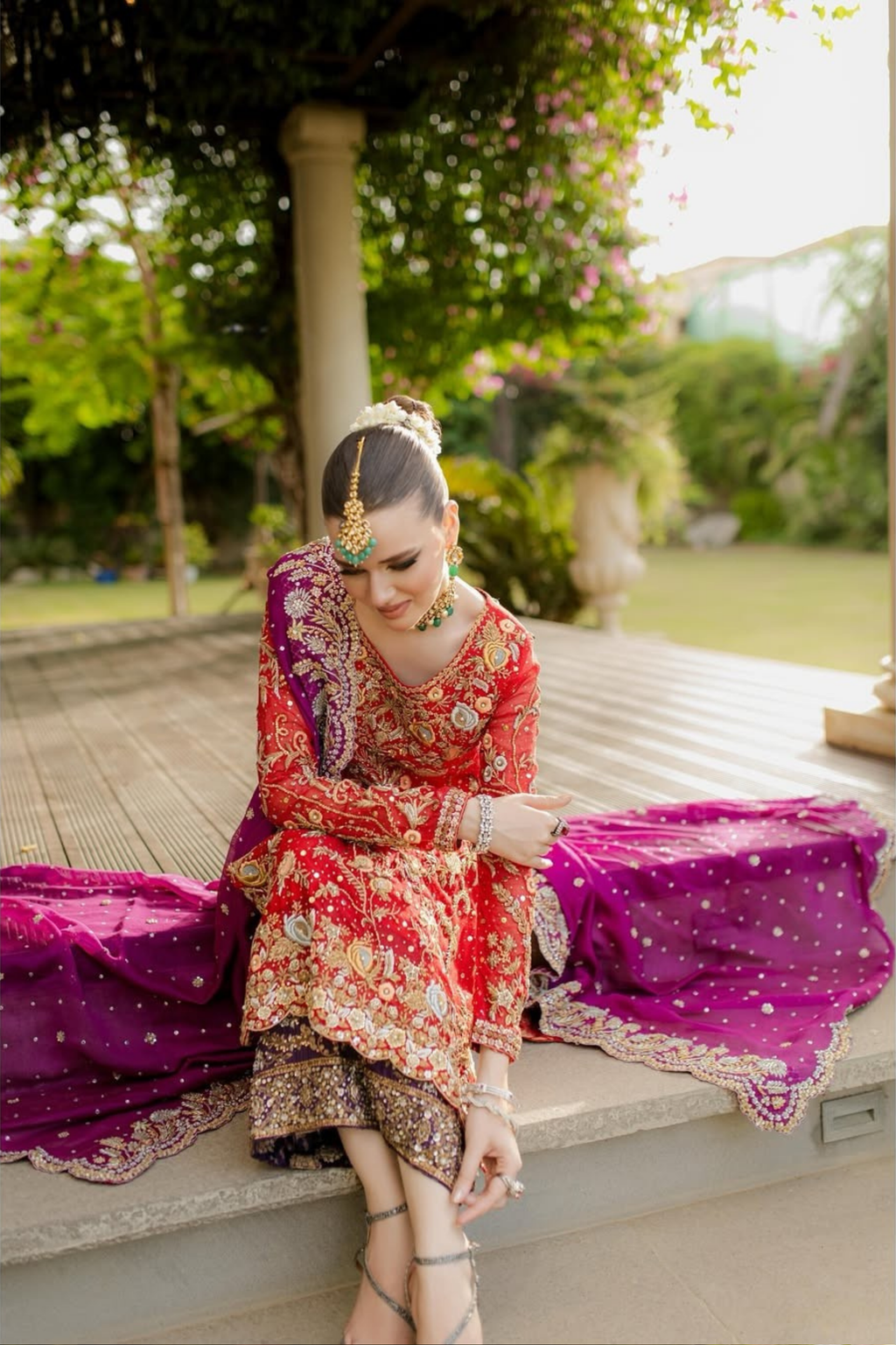 Woman in traditional red and gold outfit with purple dupatta sitting outdoors.
