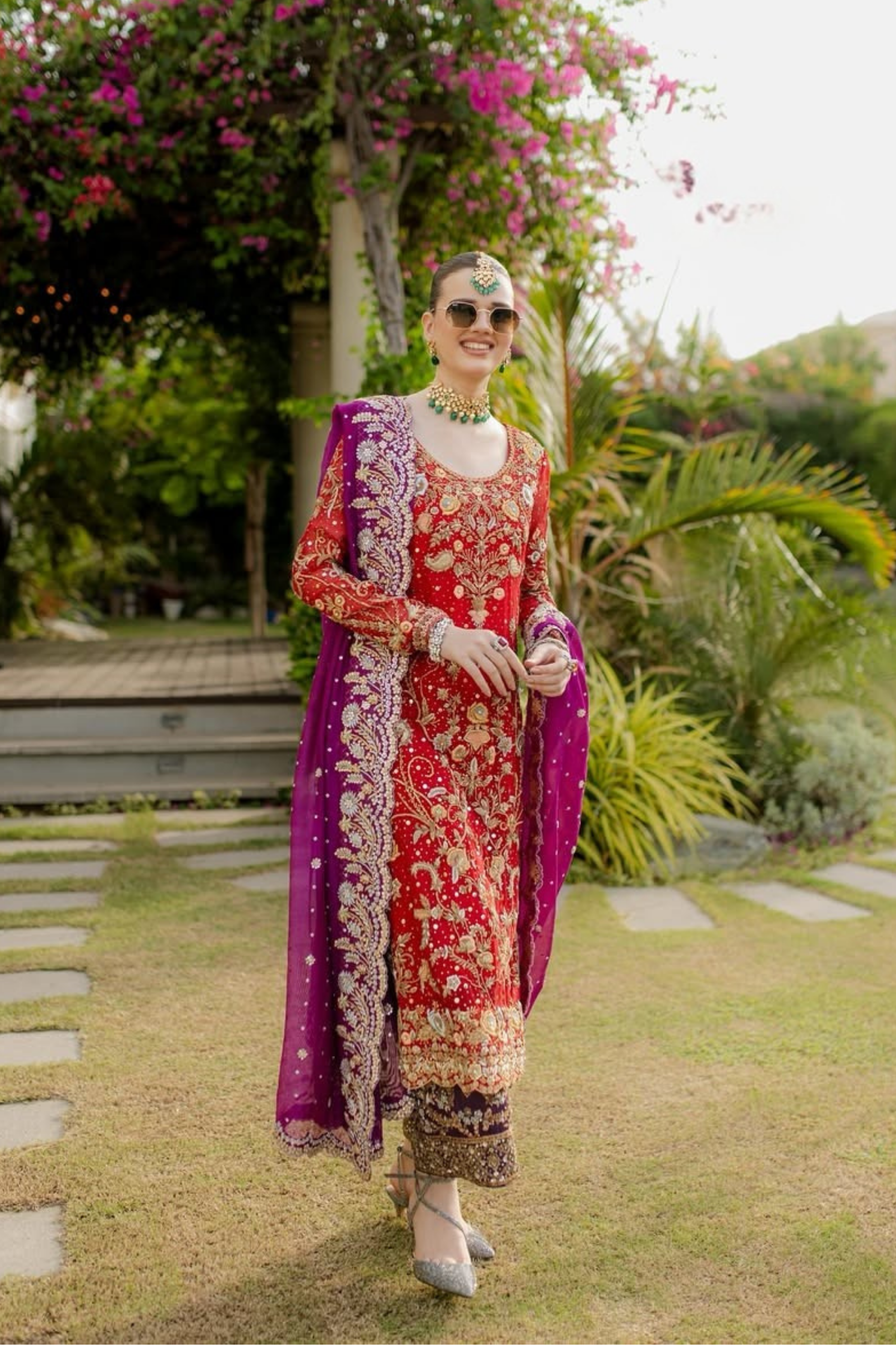 Woman in a colorful traditional outfit standing in a garden with greenery and flowers.