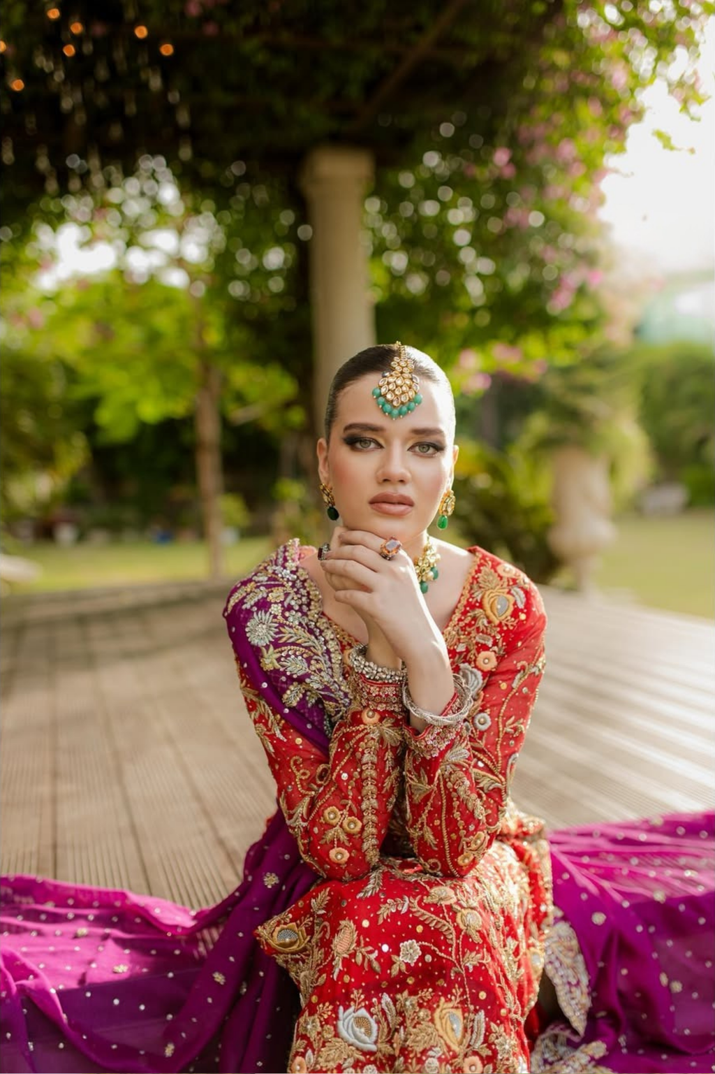 Woman in traditional red and purple outfit with jewelry, sitting outdoors.
