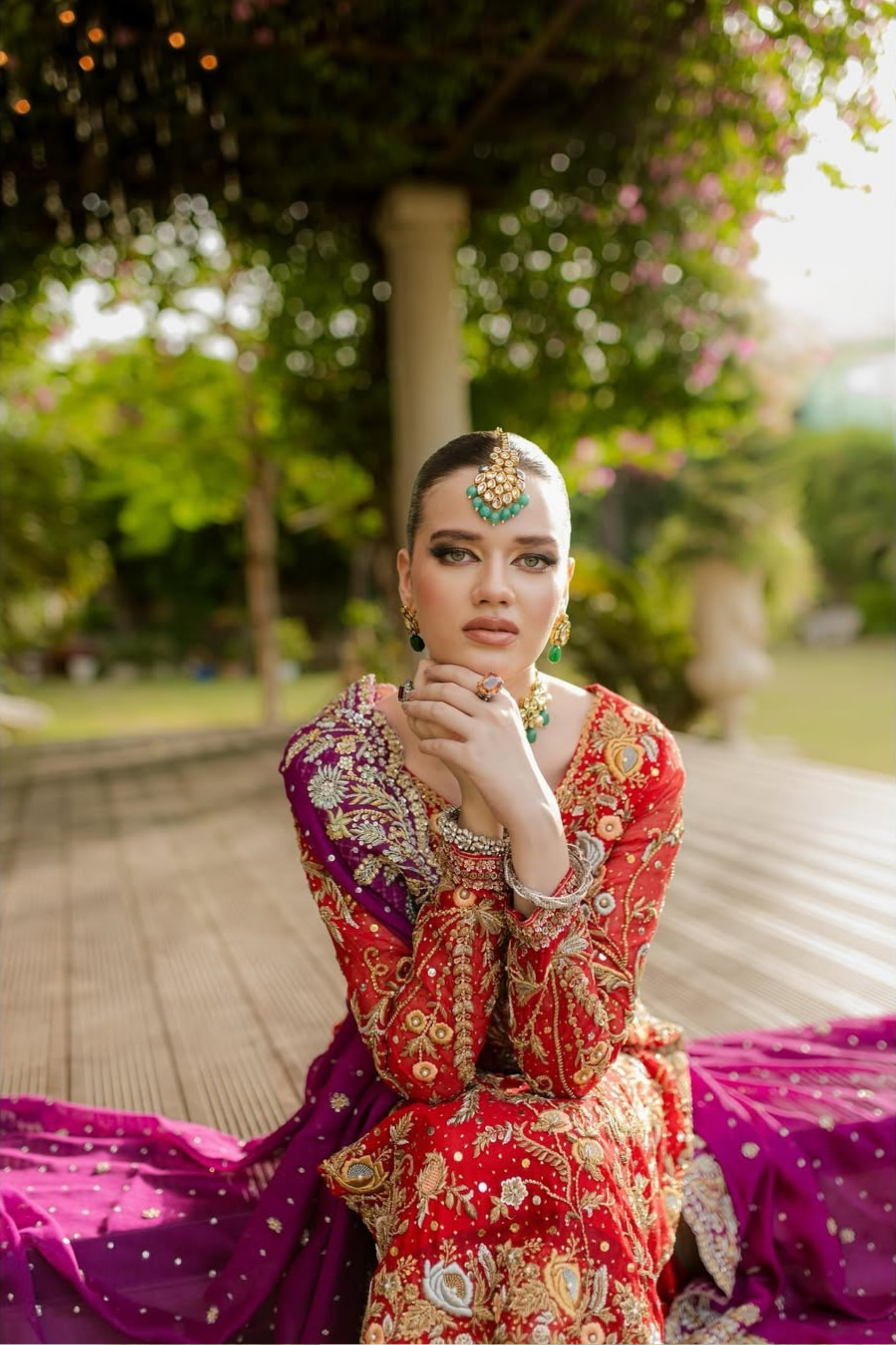 Woman in traditional red and purple outfit with jewelry, sitting outdoors.