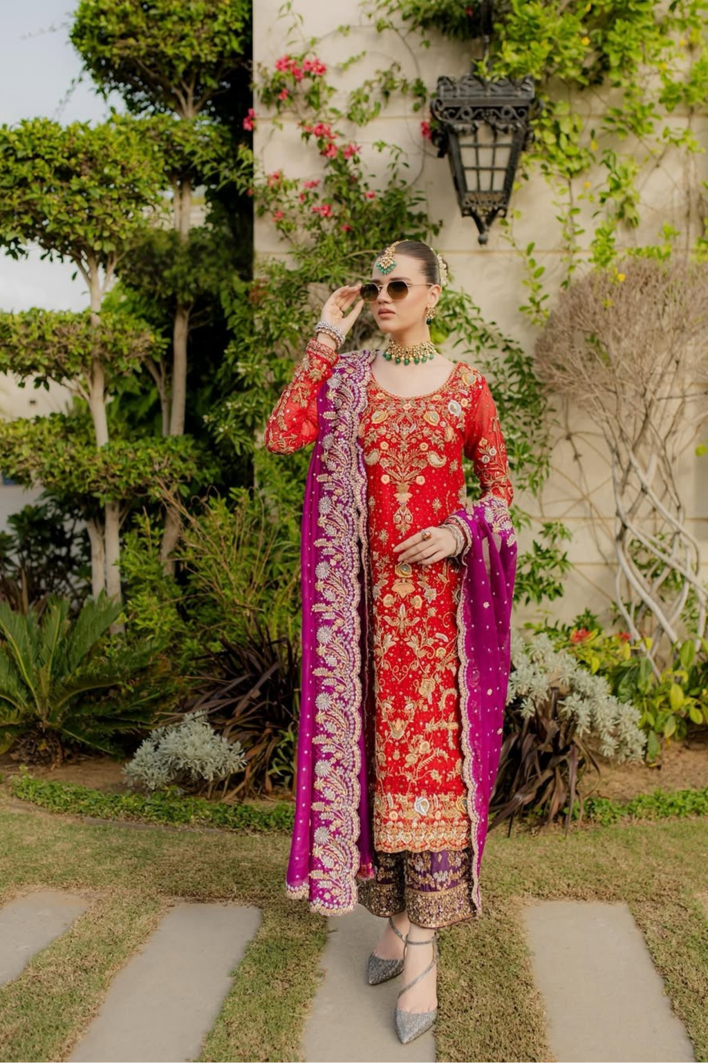 Woman in traditional red and pink embroidered outfit standing outdoors with greenery.