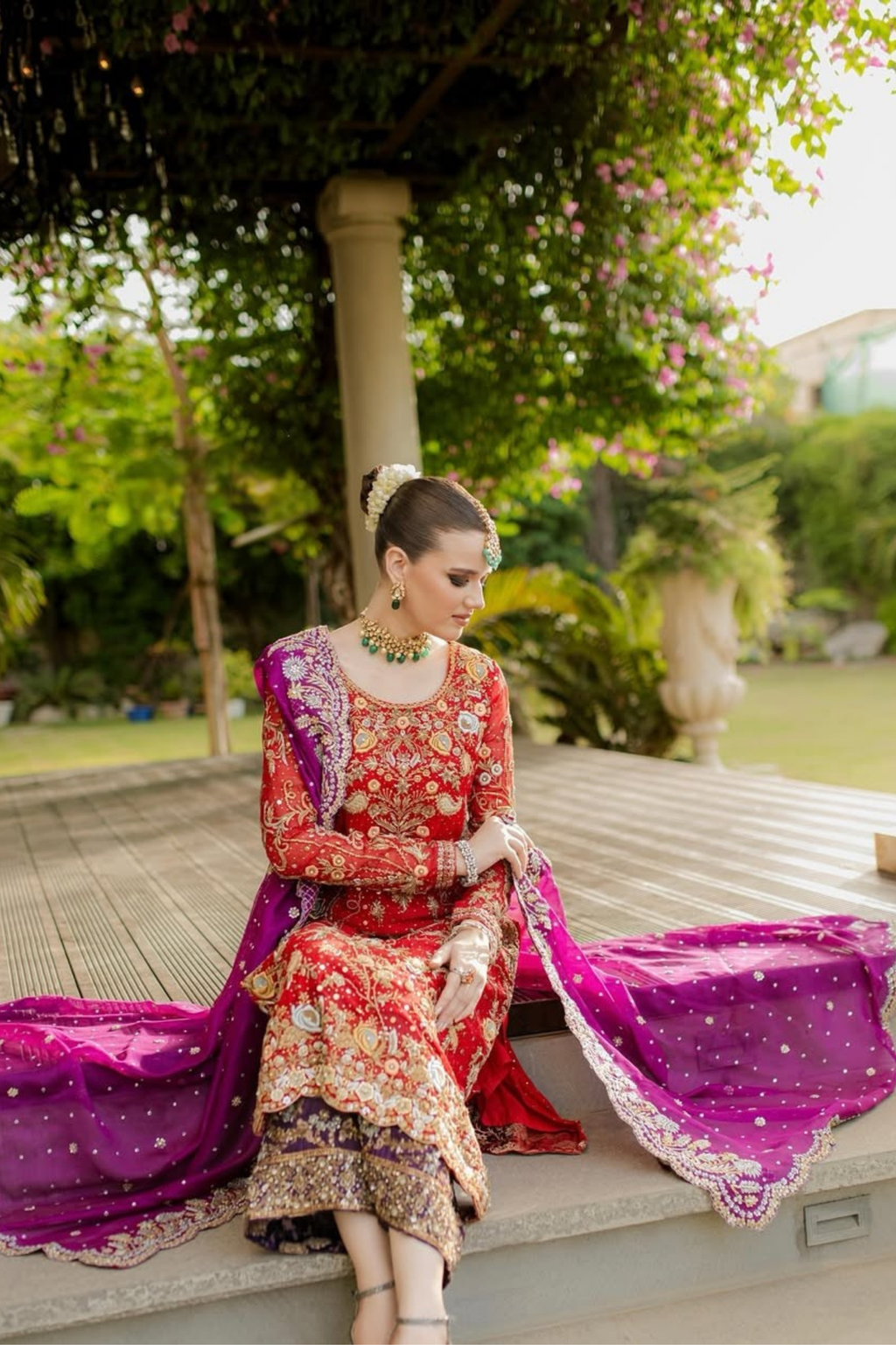 Woman in traditional red and purple outfit sitting outdoors with greenery in the background
