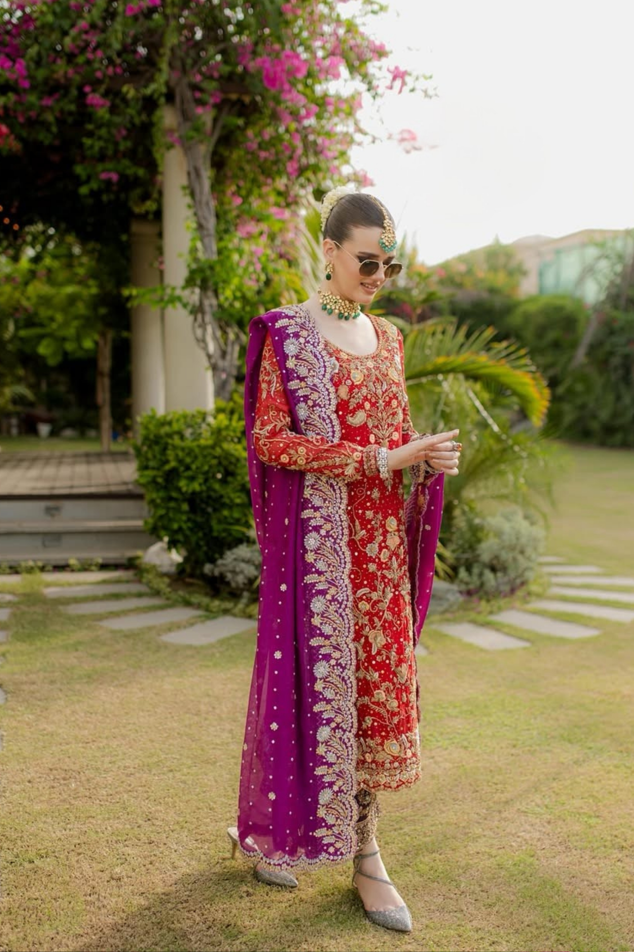 Woman in a red and purple traditional outfit standing outdoors with greenery in the background