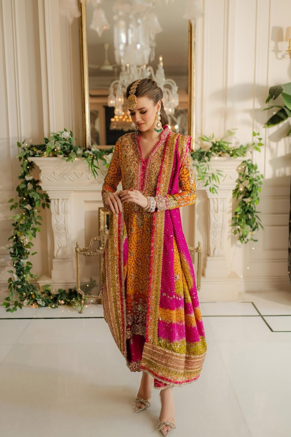 Woman in a colorful traditional outfit standing in a decorated room with greenery and chandeliers.