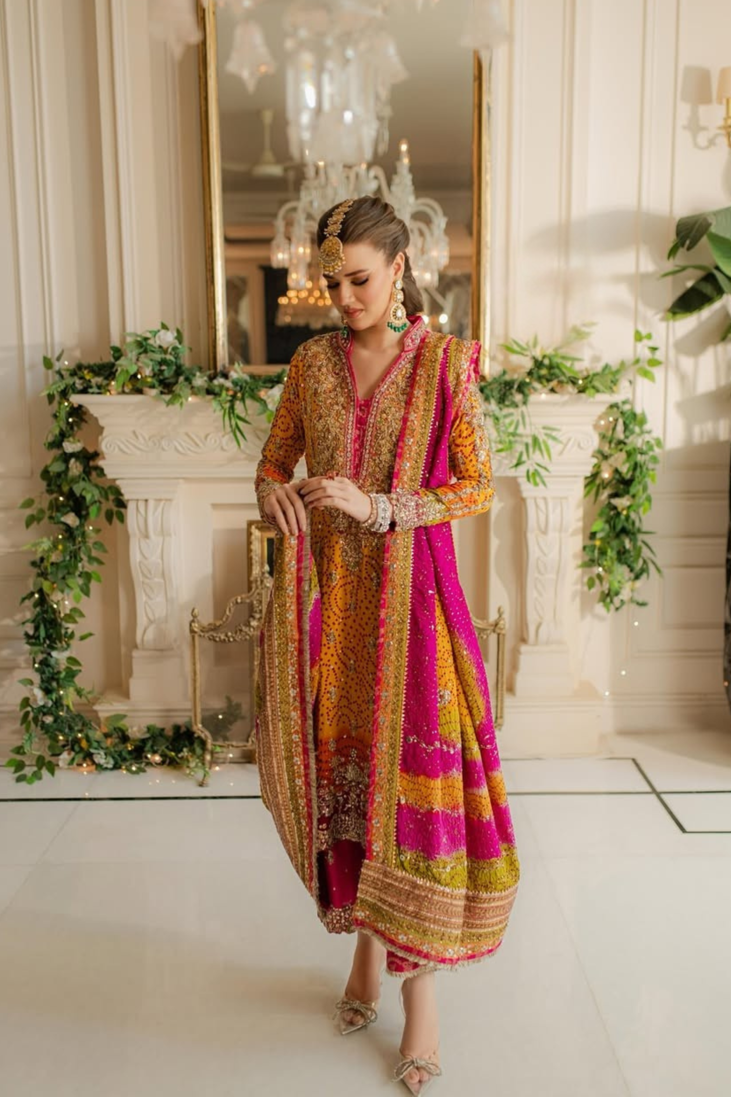 Woman in a colorful traditional outfit standing in a decorated room with greenery and chandeliers.