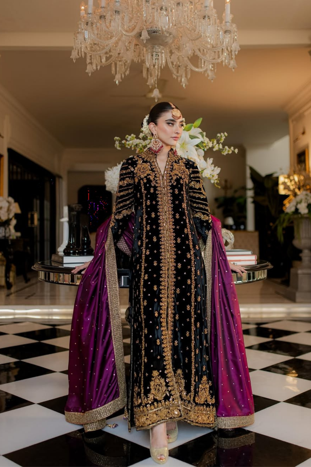 Woman in a black and gold embroidered outfit with a purple dupatta standing in an elegant interior setting.