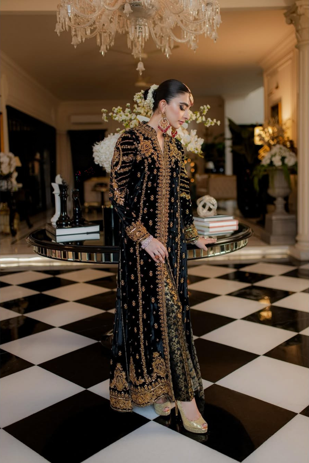 Woman in an ornate black and gold dress standing in a room with a checkered floor and chandelier.