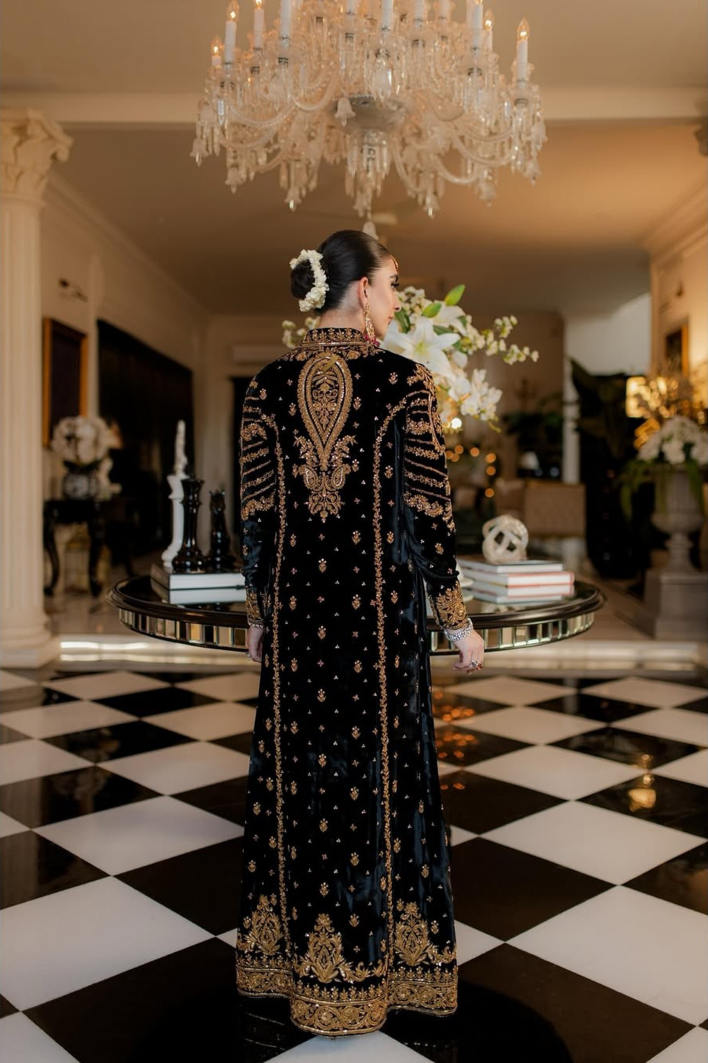 Woman in an ornate black and gold dress standing in a room with a chandelier and checkered floor.