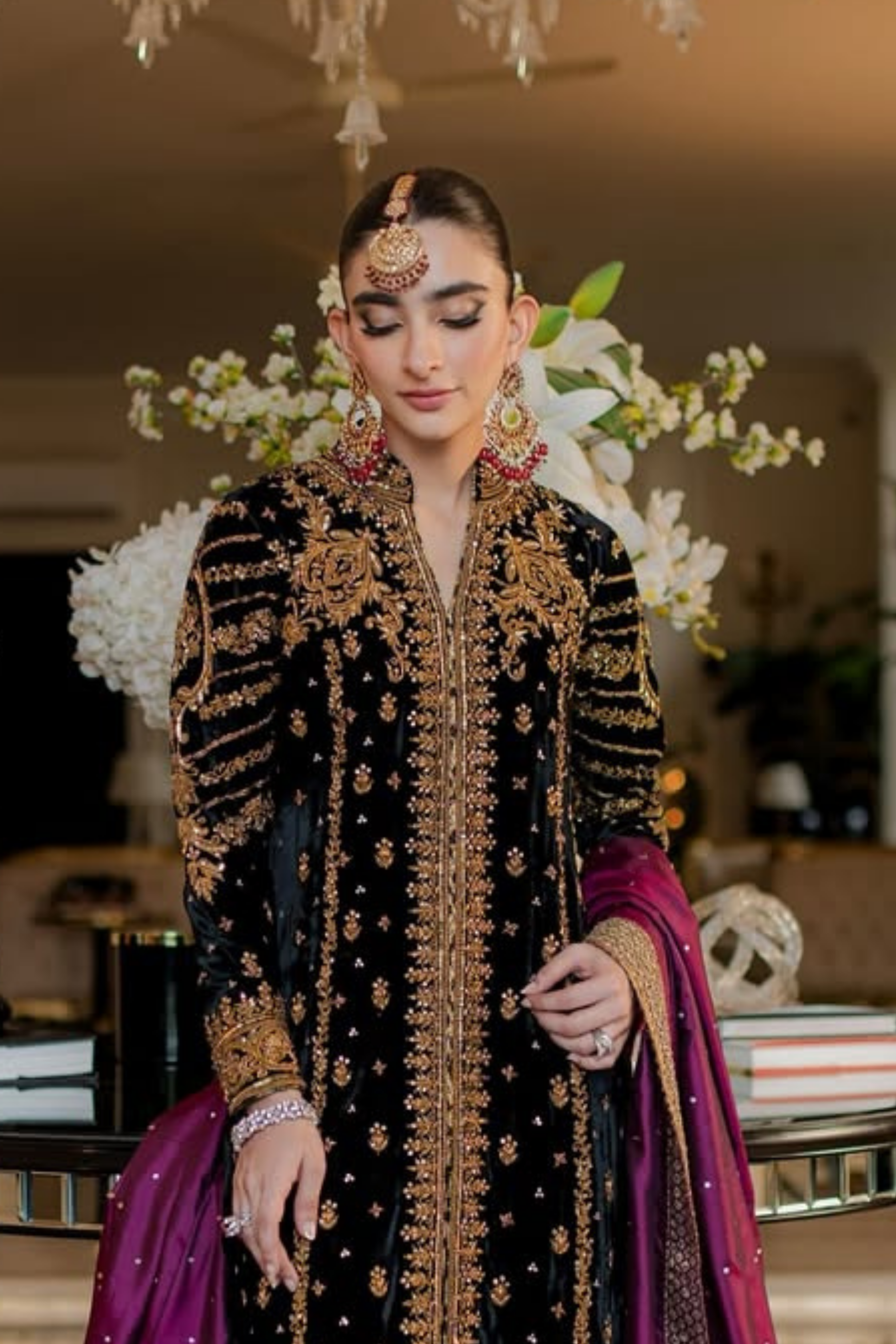Woman in traditional black and gold embroidered outfit with purple dupatta, indoor setting.
