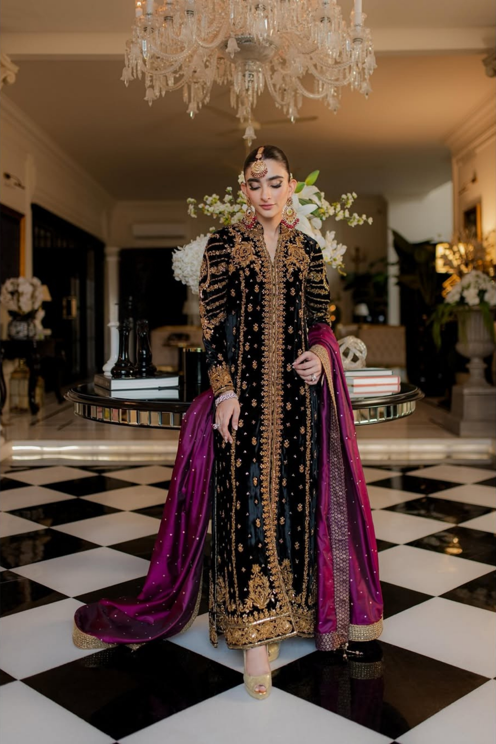 Woman in an ornate black and gold outfit with a purple dupatta standing in a decorated room.