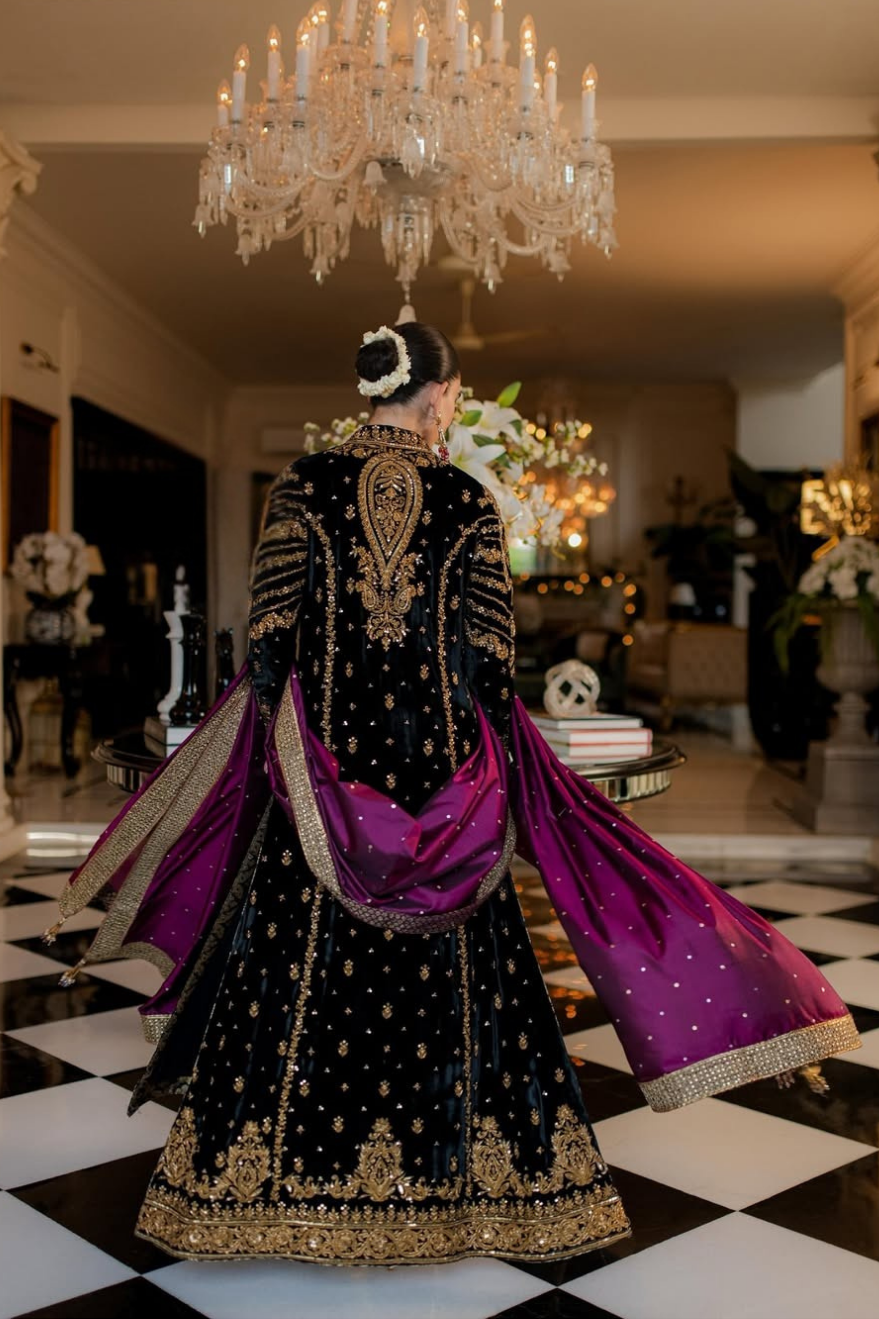 Person wearing a black and gold embroidered outfit with a purple dupatta in an elegant room with chandeliers.