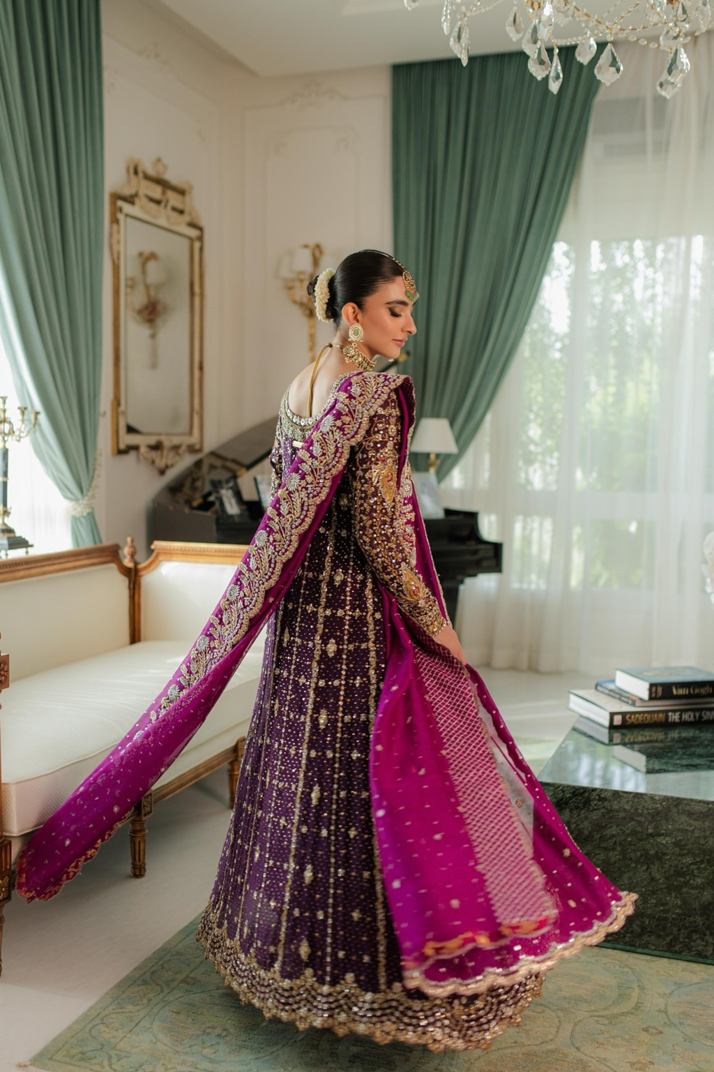 Woman in traditional embroidered outfit in a decorated room with green curtains and a chandelier.
