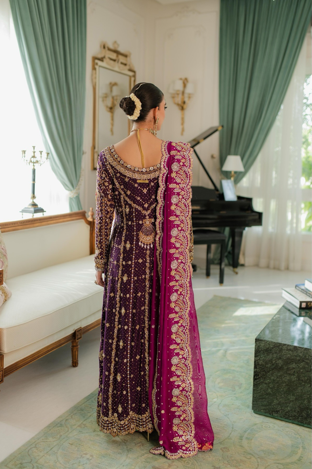 Woman in a traditional purple and pink saree standing in a elegant room with a piano and green curtains.