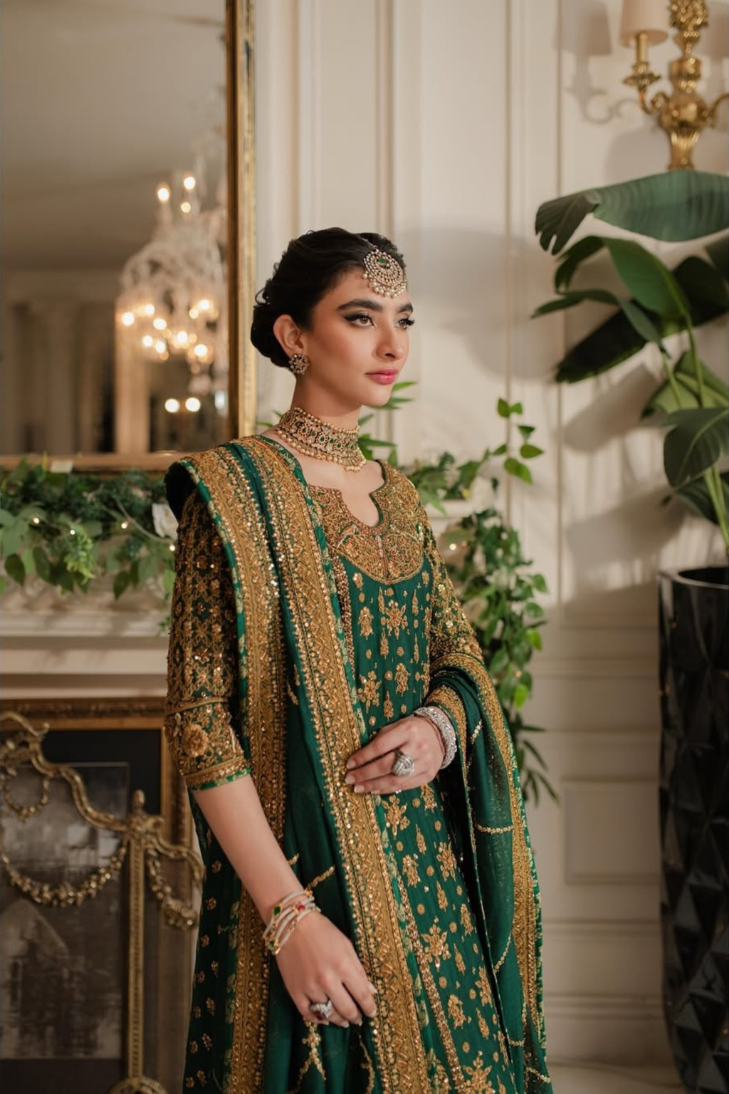 Woman in a green and gold traditional outfit with jewelry in an elegant indoor setting.