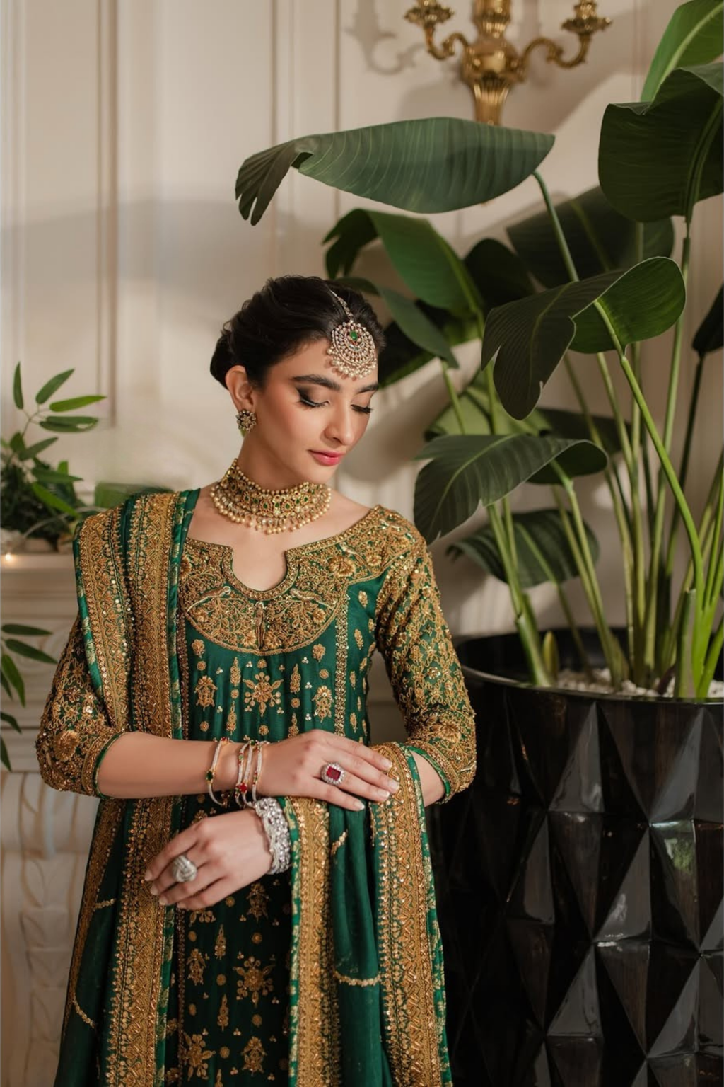 Woman in traditional green and gold outfit with jewelry, standing indoors with plants in the background