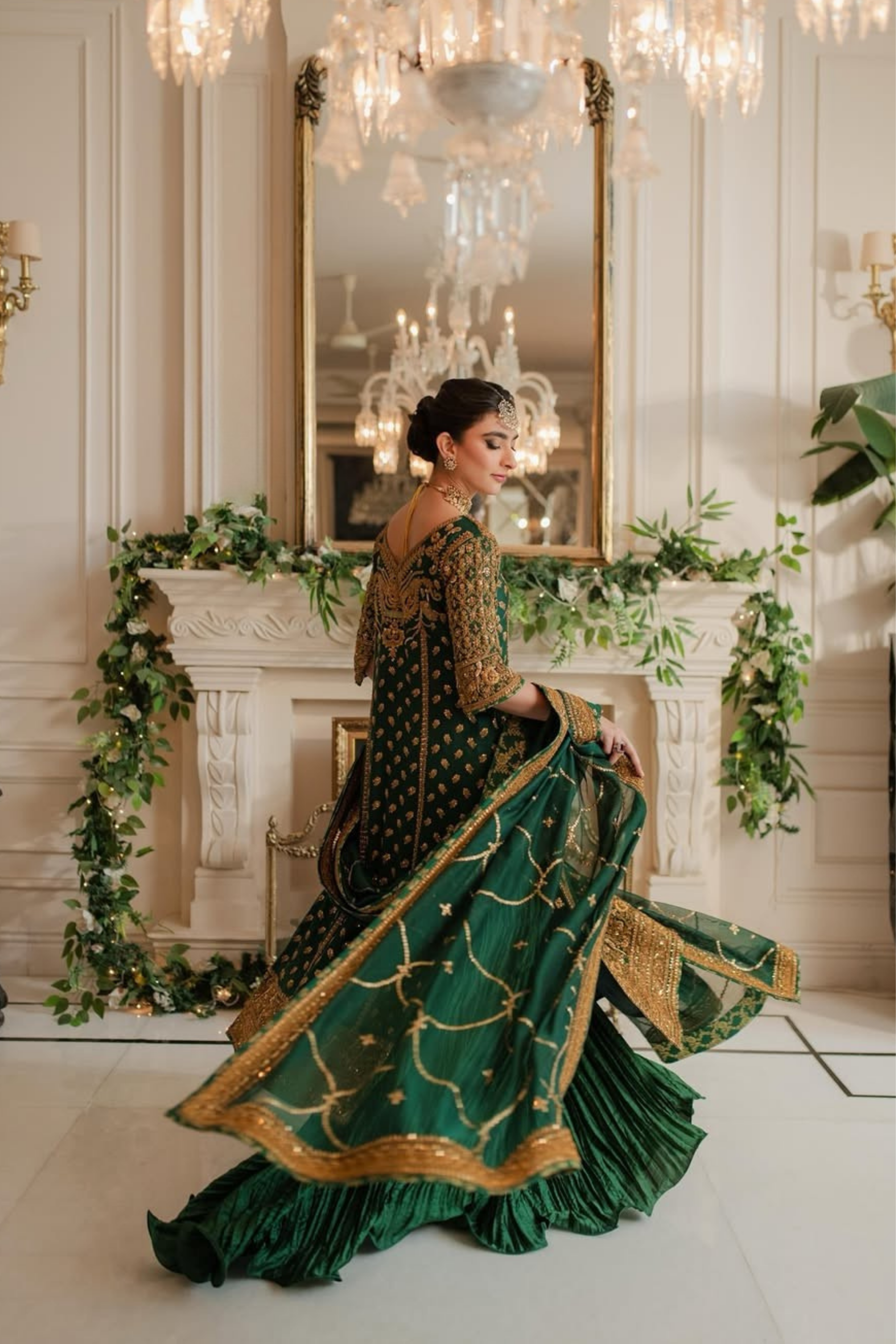 Woman in a green and gold traditional outfit standing in a decorated room with chandeliers and greenery.