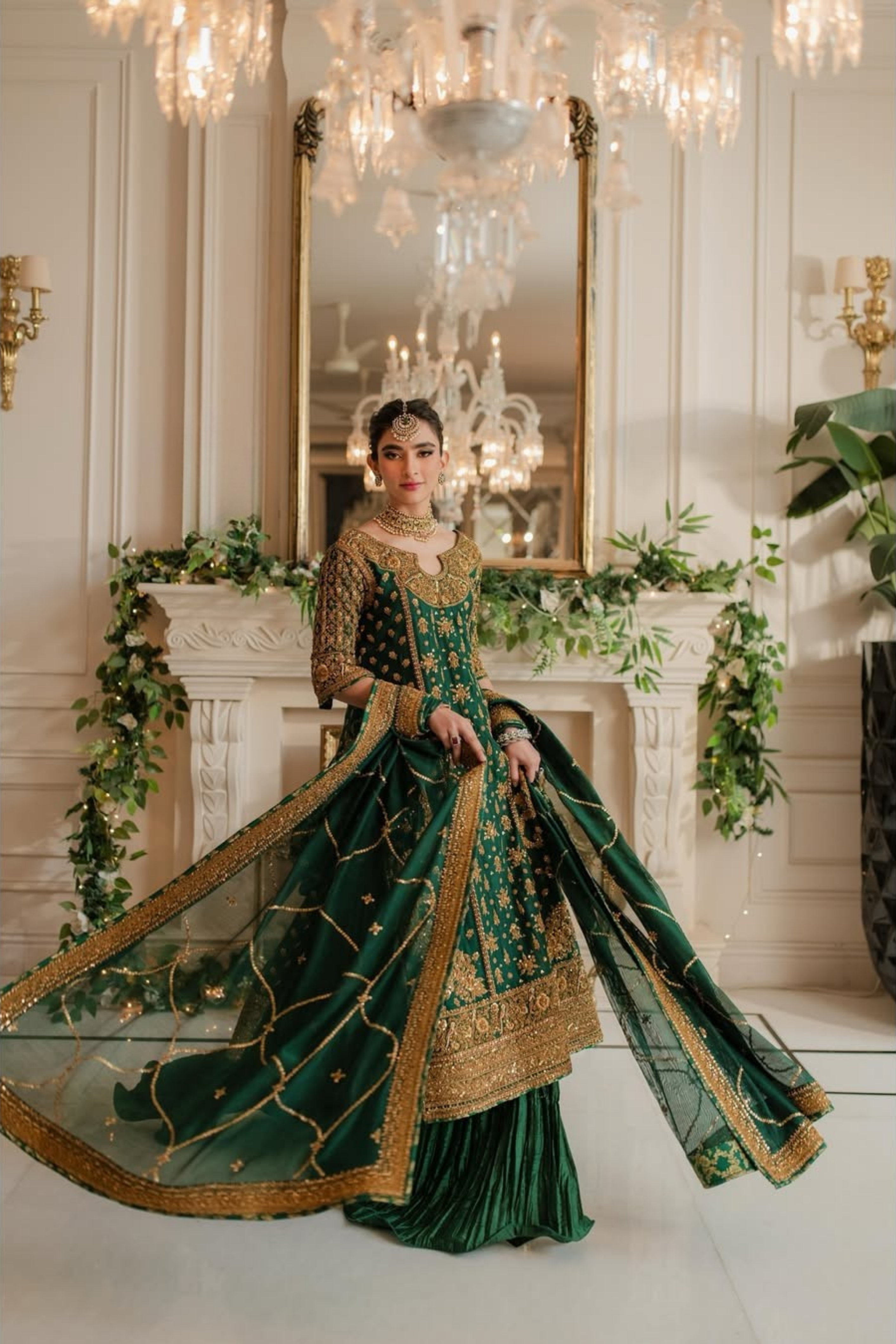 Woman in a green and gold traditional outfit standing in a decorated room with chandeliers and floral arrangements.