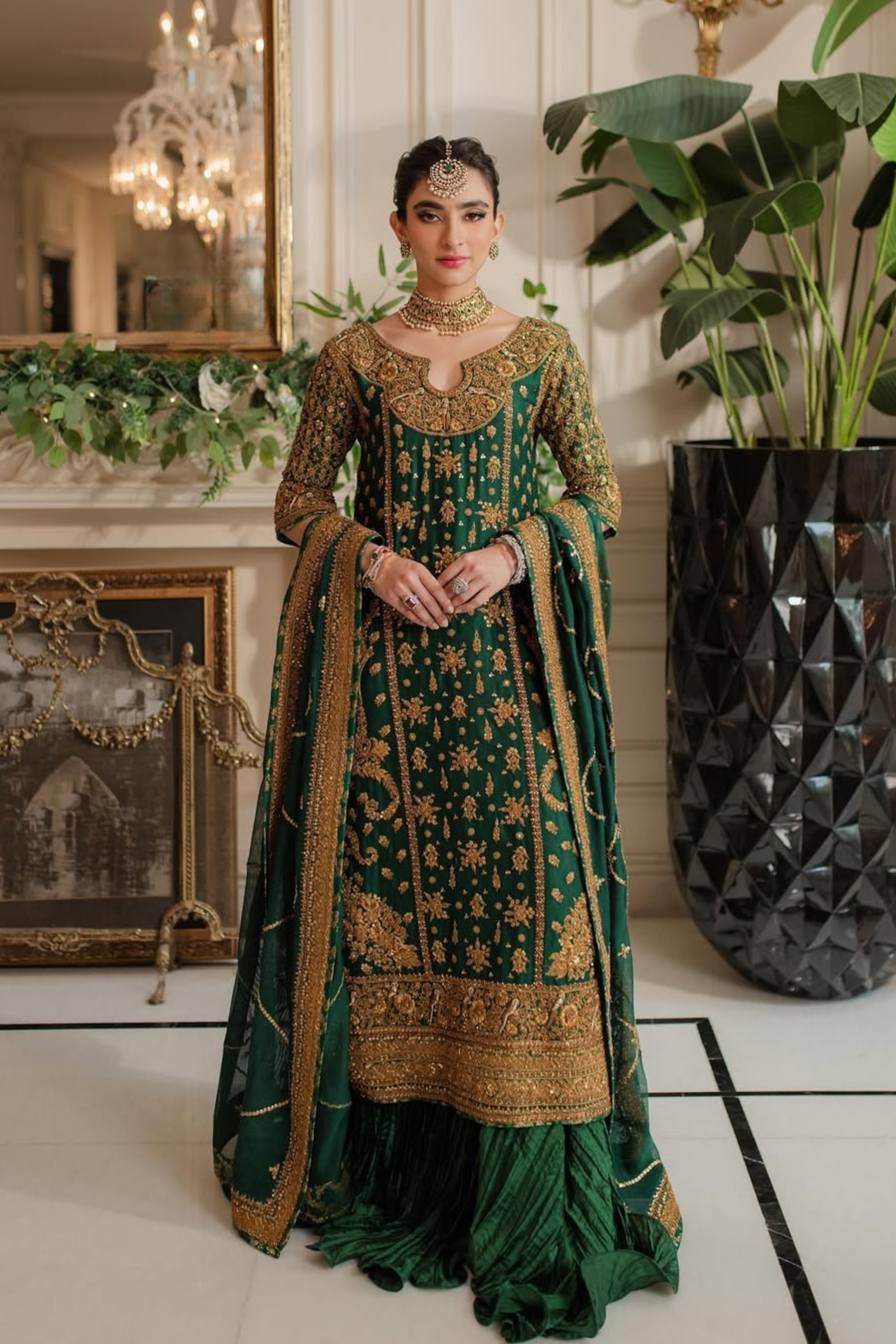 Woman in a green and gold traditional outfit standing in an elegant room with plants and decorative items.