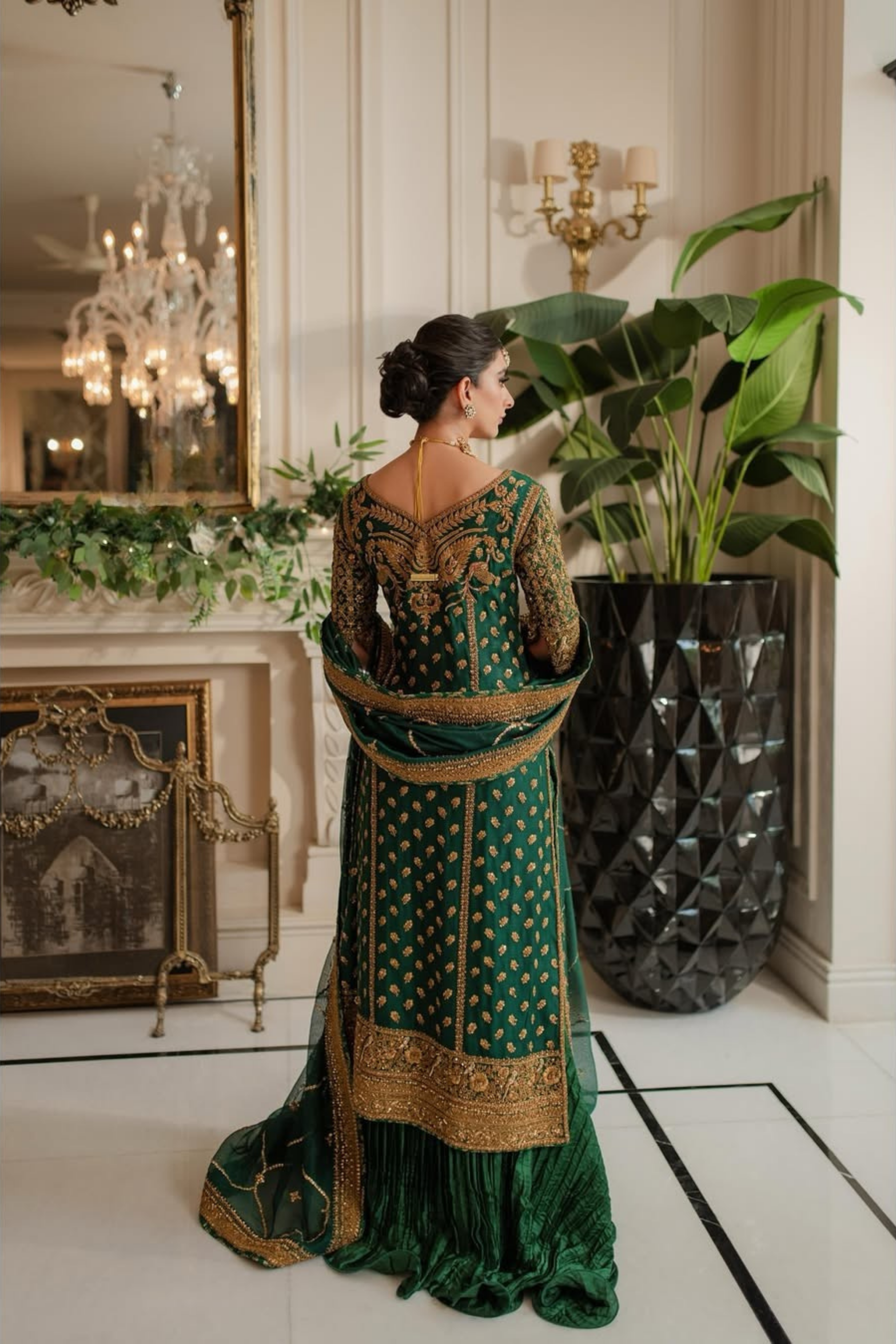 Woman in a green and gold traditional outfit standing in an elegant room with chandeliers and plants.