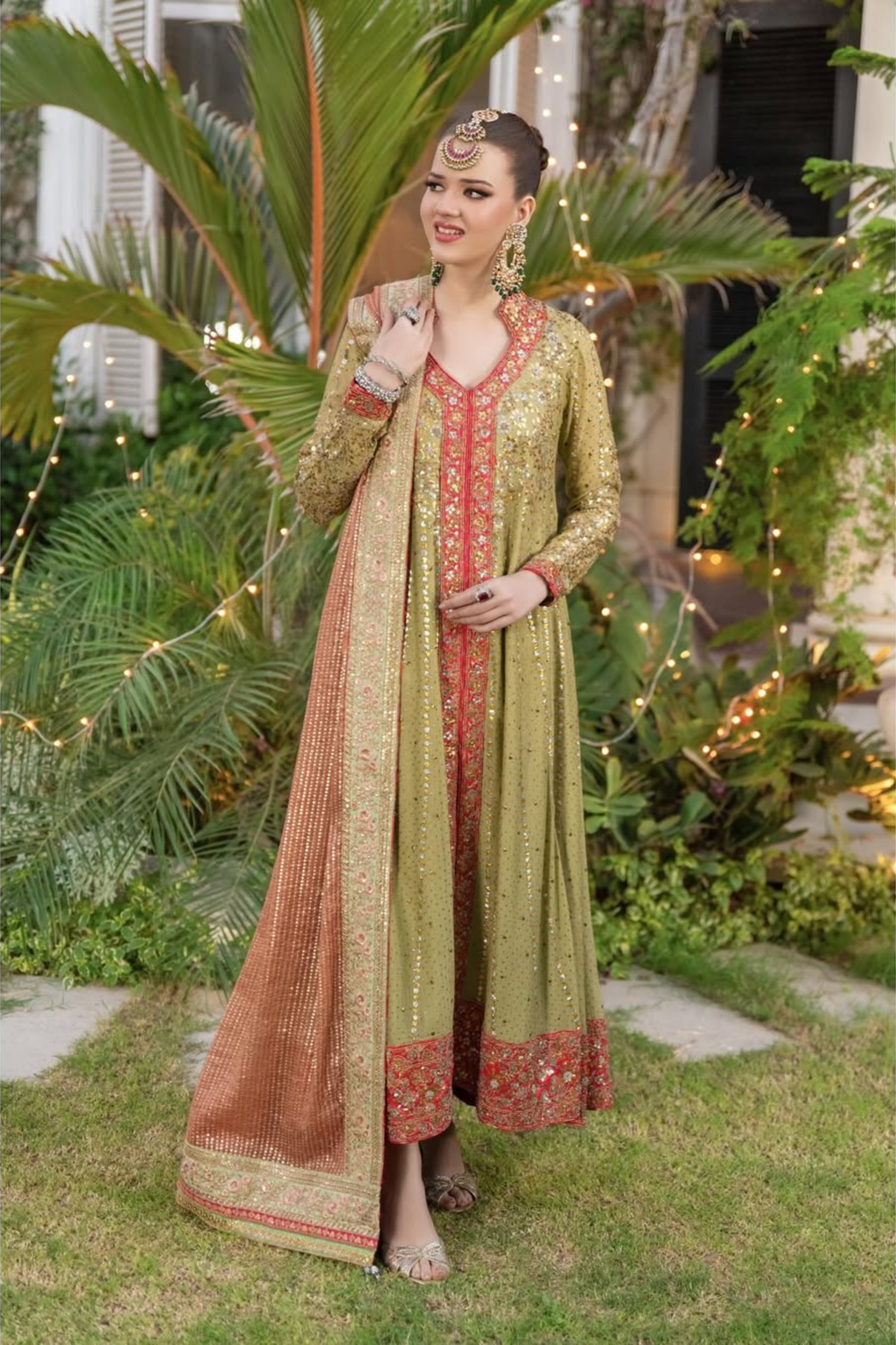 Woman in a green and red traditional outfit standing outdoors with plants and lights in the background