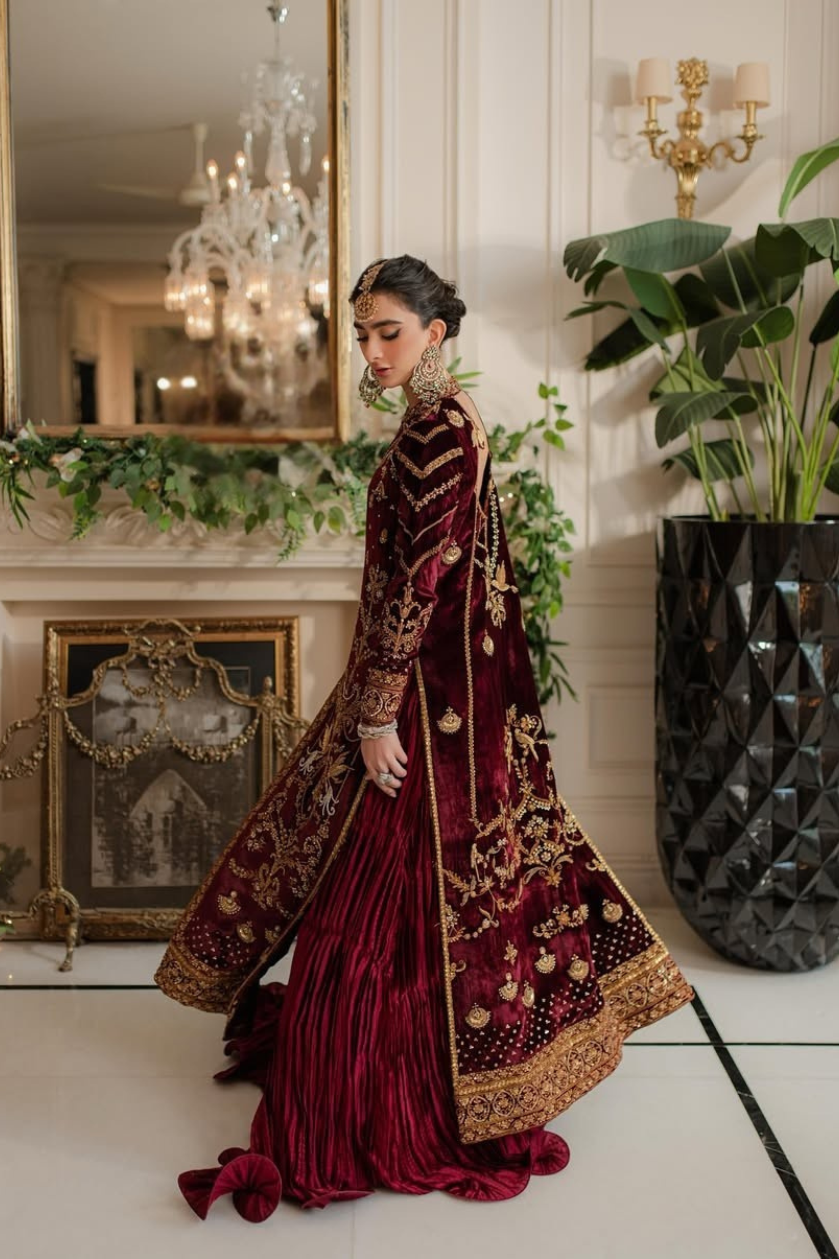 Woman in a burgundy and gold traditional outfit standing in an elegant room with chandelier and plants.