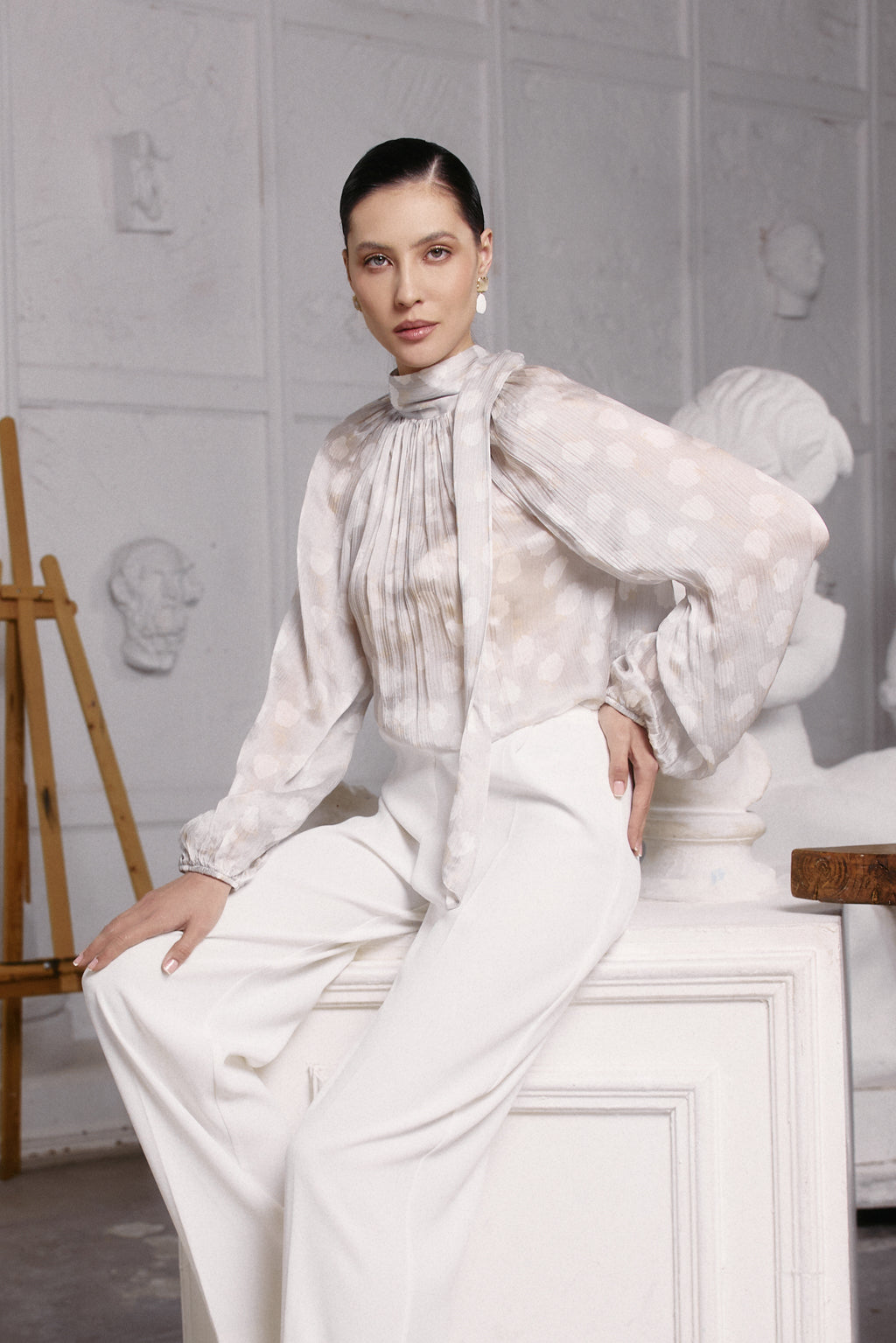 Woman in a white outfit sitting on a white pedestal in a minimalistic setting.