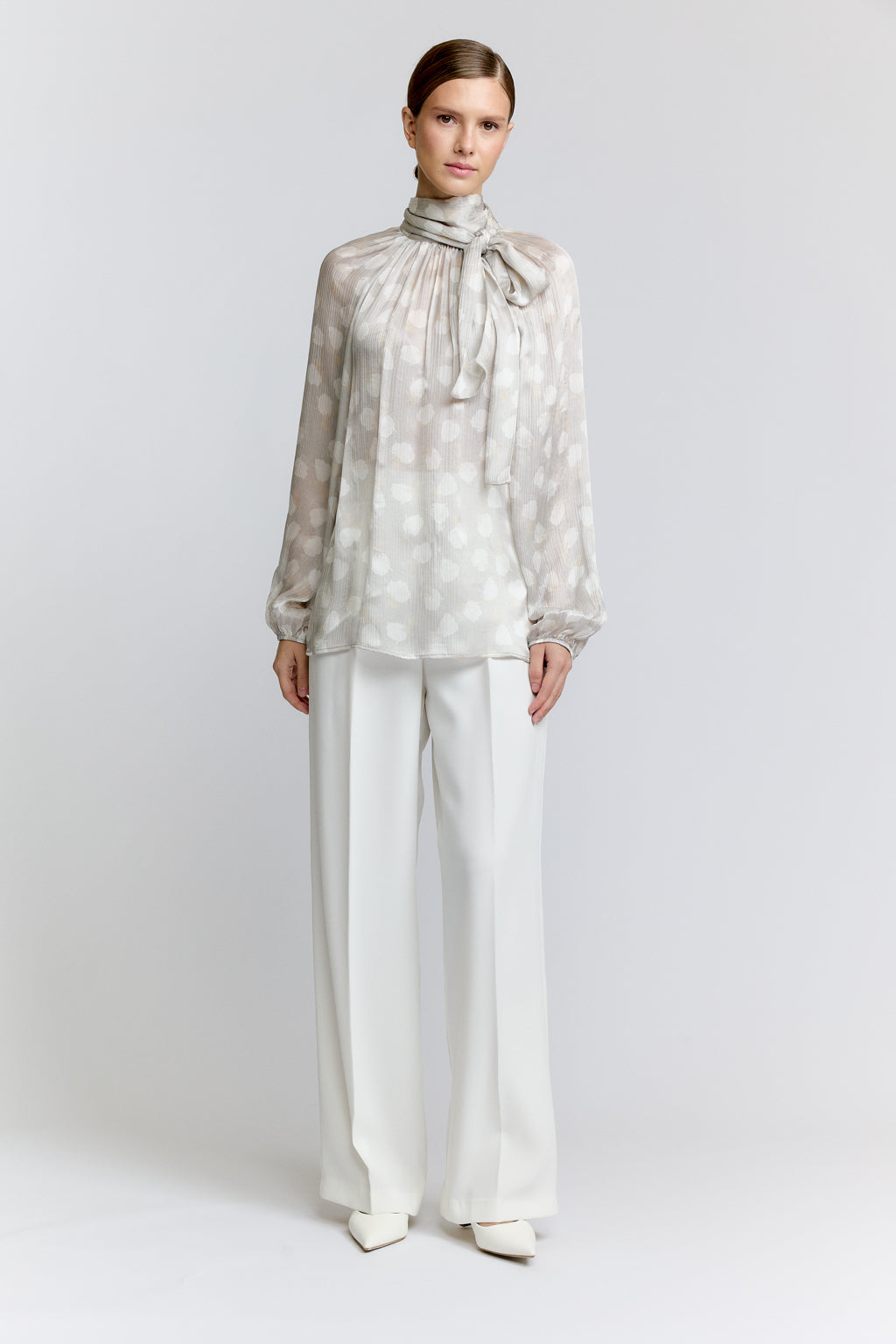 Woman wearing a patterned blouse with a bow and white pants on a plain background