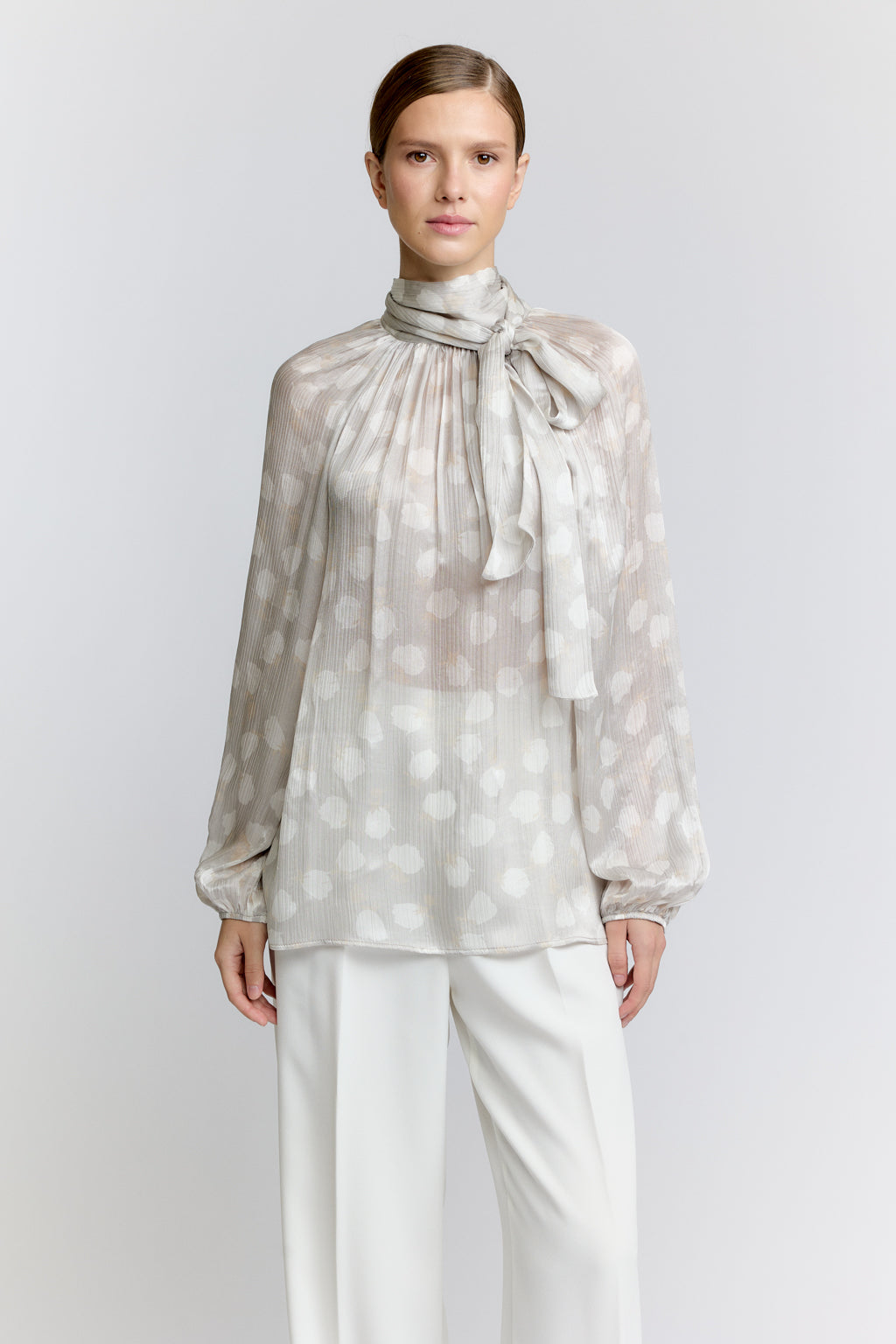 Woman wearing a sheer polka dot blouse with a tie and white pants on a light gray background