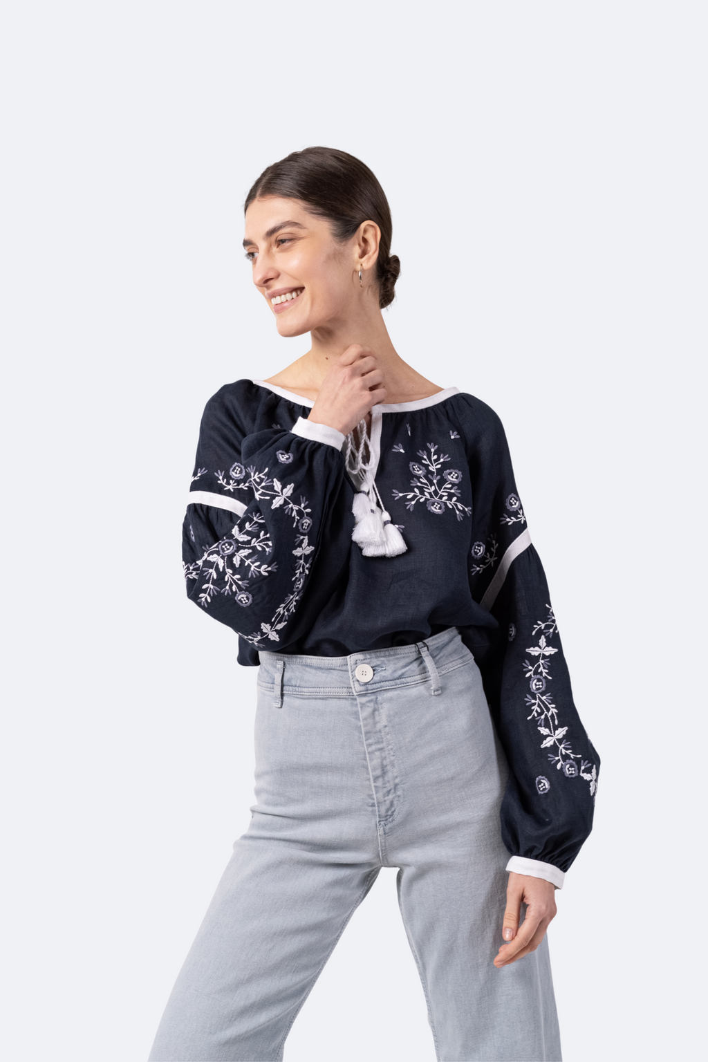 Woman wearing a navy blue blouse with white floral embroidery and light gray pants on a white background