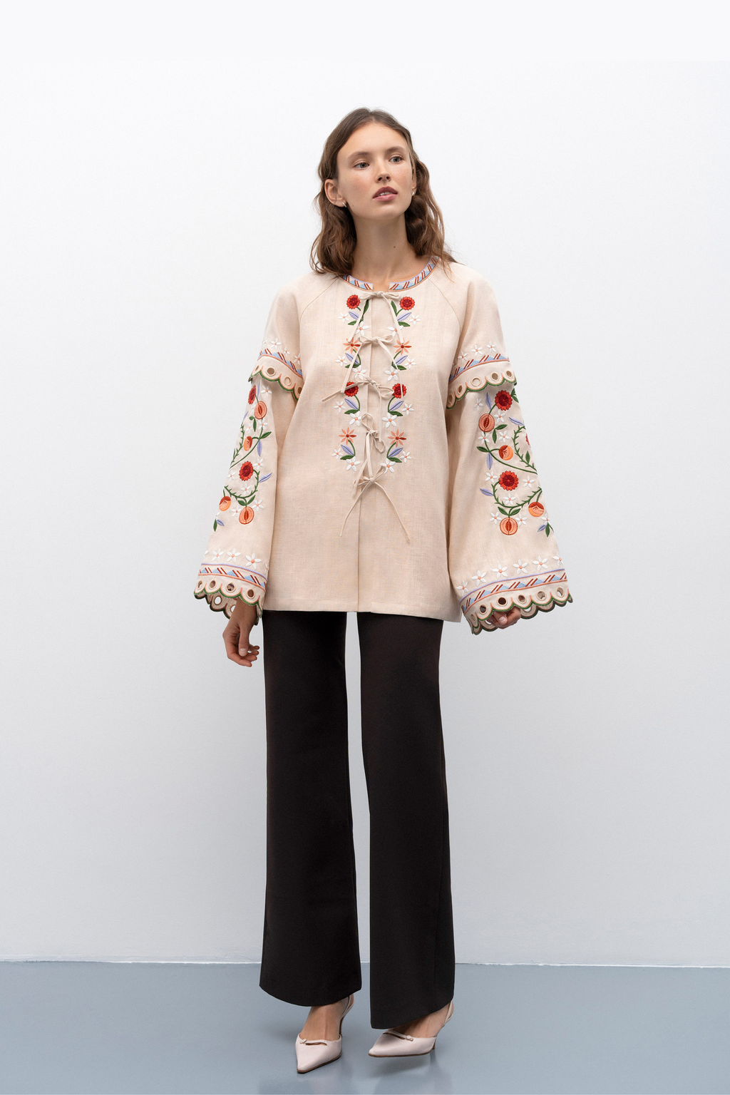 Woman wearing a beige blouse with floral embroidery and black pants on a white background