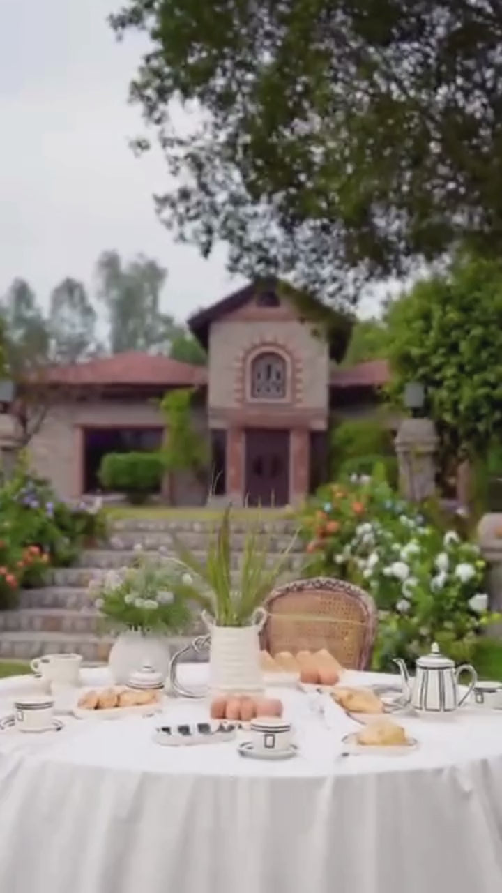 Woman in a pink traditional outfit standing outdoors near a table with a garden and building in the background.