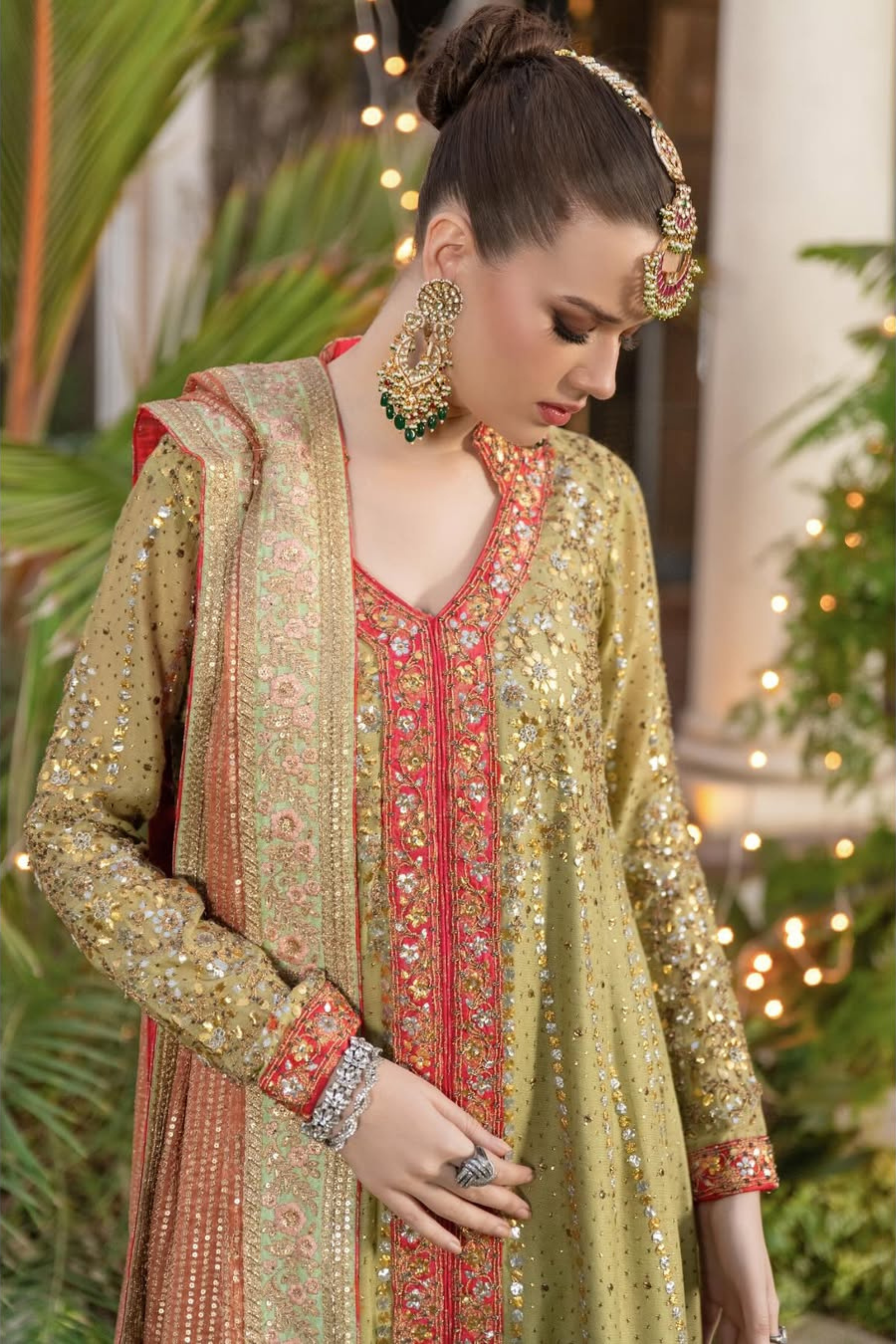 Woman in traditional embroidered outfit sitting outdoors with greenery and lights in the background