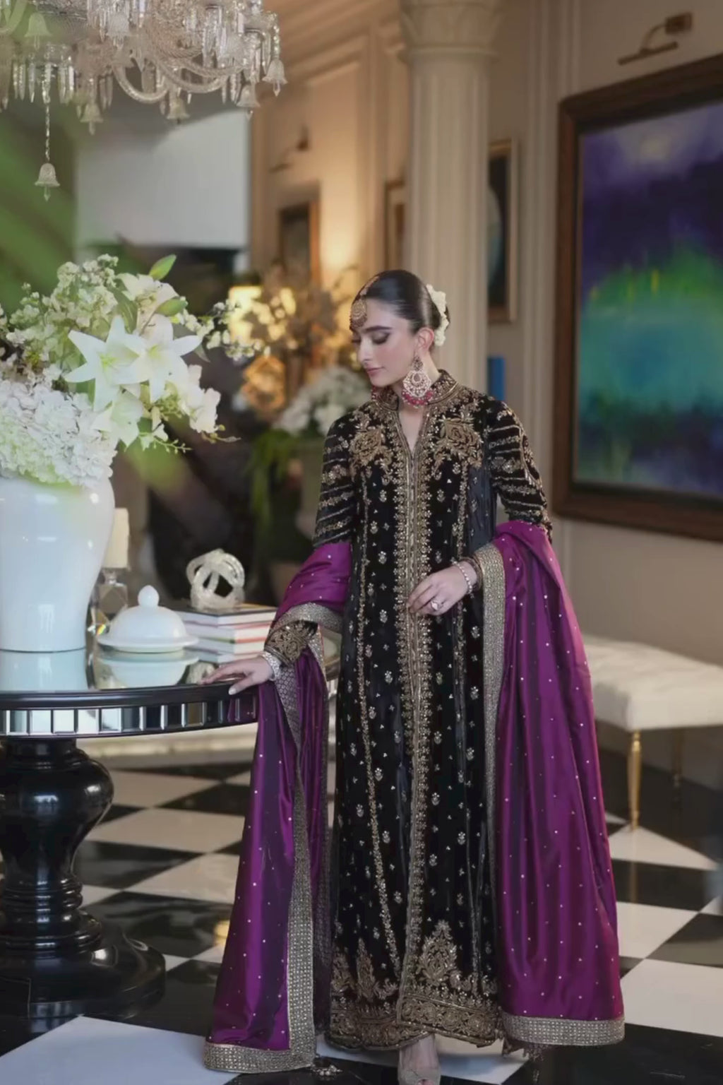 Woman in traditional black and gold embroidered outfit with purple dupatta, indoor setting.