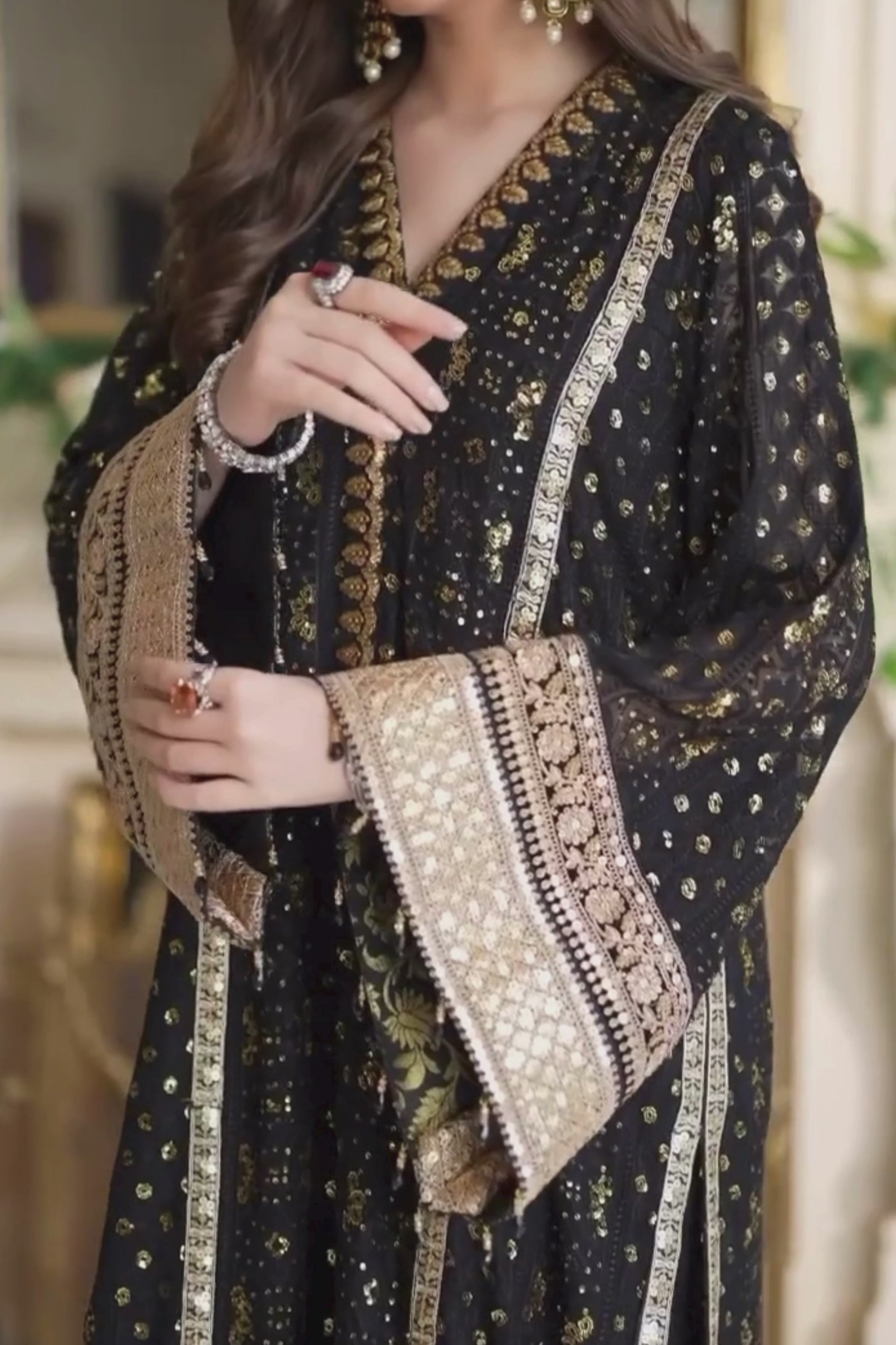 Woman in a black and gold traditional outfit standing in an elegant room with chandeliers and greenery.